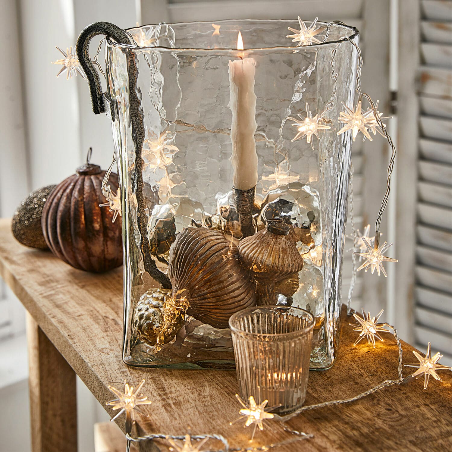 Lichtsnoer Nelisse Glazen windlicht met metalen handvat en koperen decoraties op houten tafel met stervormige lichtjes