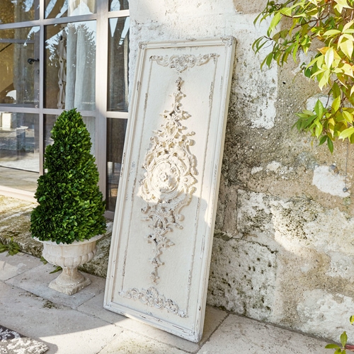 Panneau décoratif en bois blanc avec relief vintage appuyé contre un mur en pierre près d'une plante verte