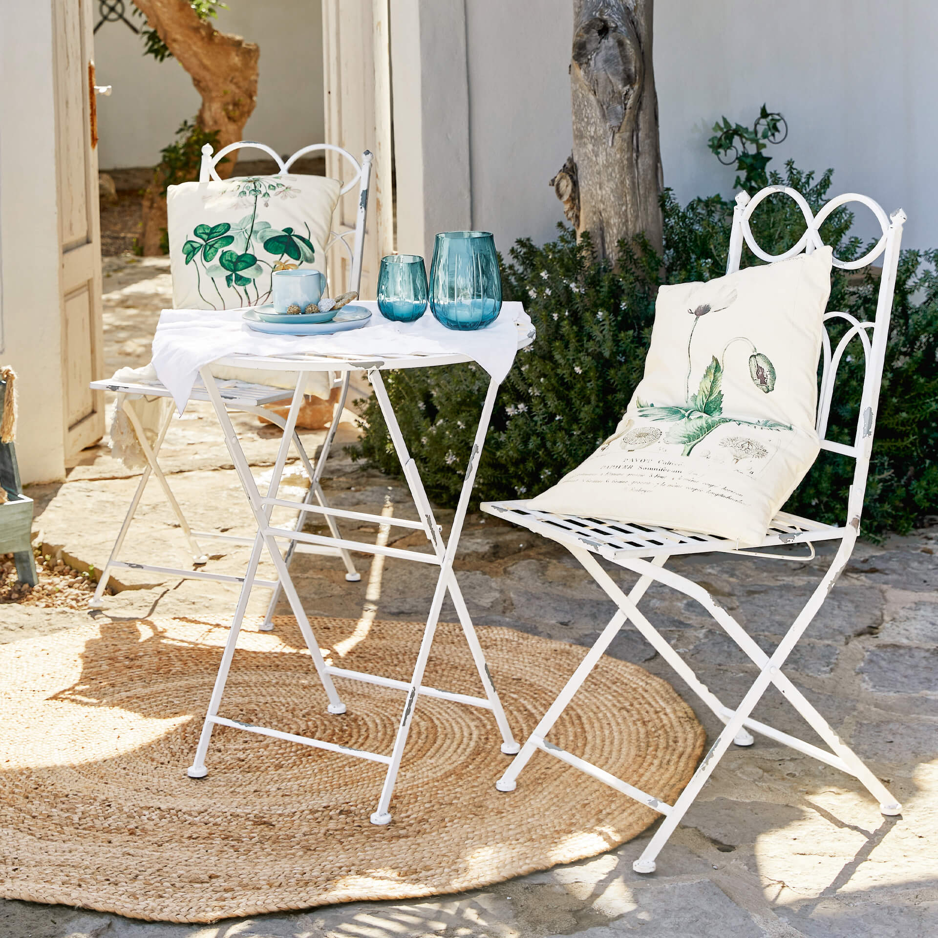 Table et chaises de jardin en métal blanc avec coussins à motifs de feuilles vertes sur terrasse en pierre avec tapis en jute