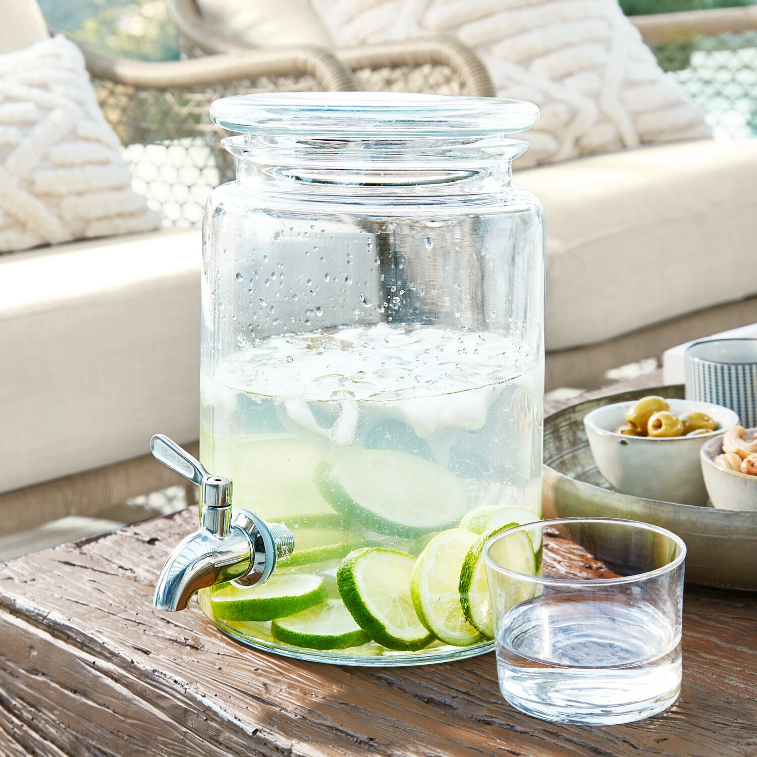 Drankdispenser Gabela Glazen drankendispenser met kraan, gevuld met water en limoenplakjes op houten tafel