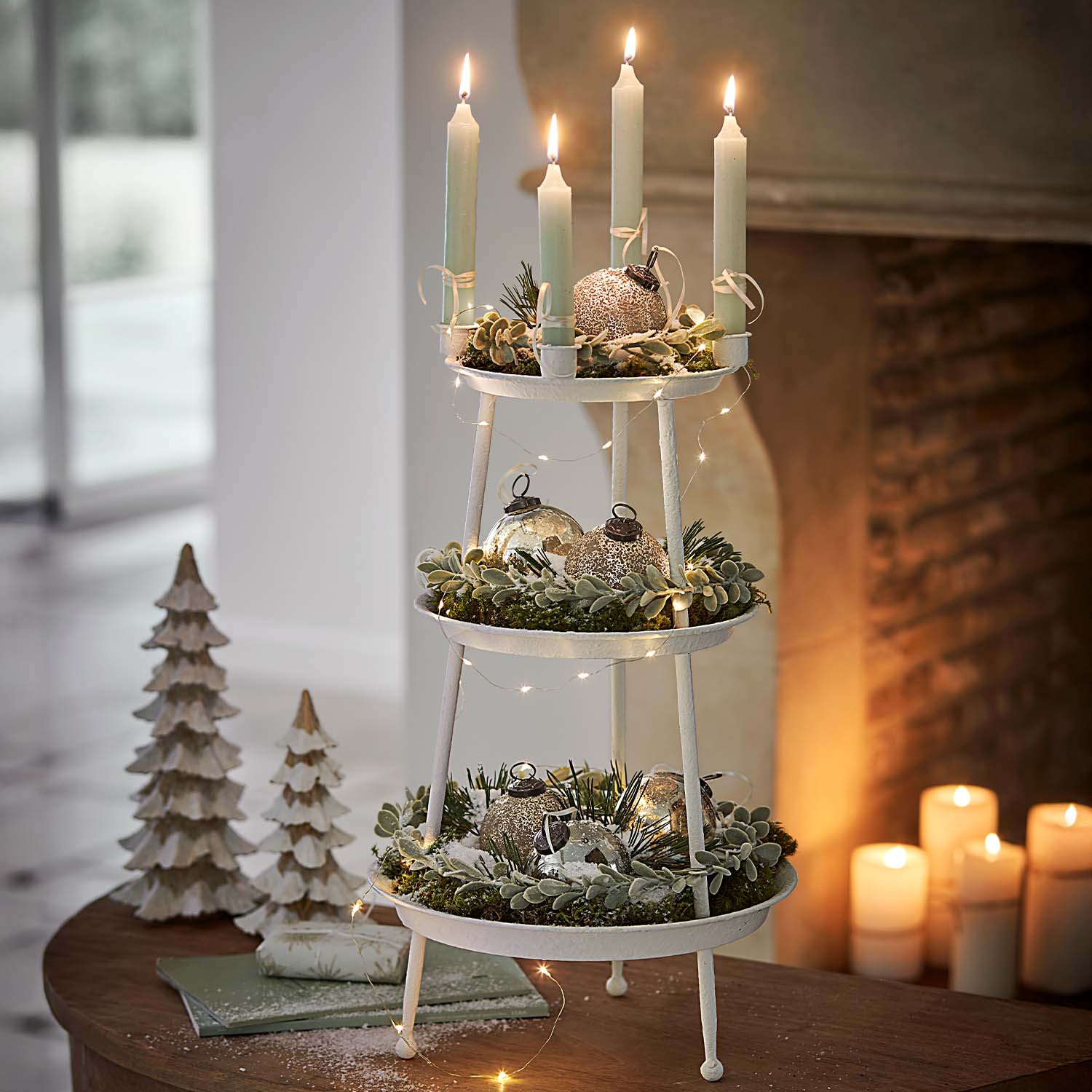 Witte metalen kerstdecoratie met drie niveaus, groene takken en brandende kaarsen op tafel