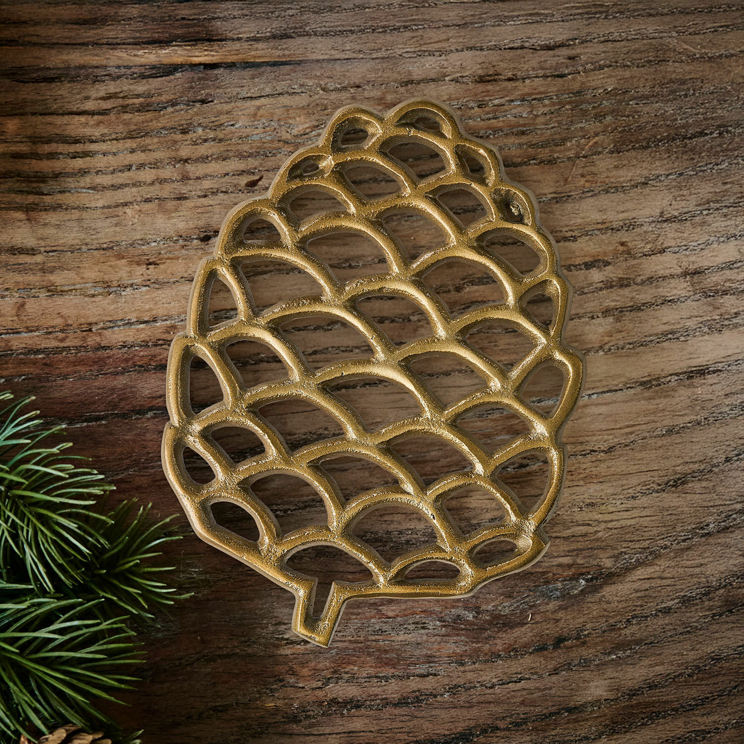Dessous de plat Caritha Dessous de plat en métal doré en forme de pomme de pin sur table en bois avec branches vertes