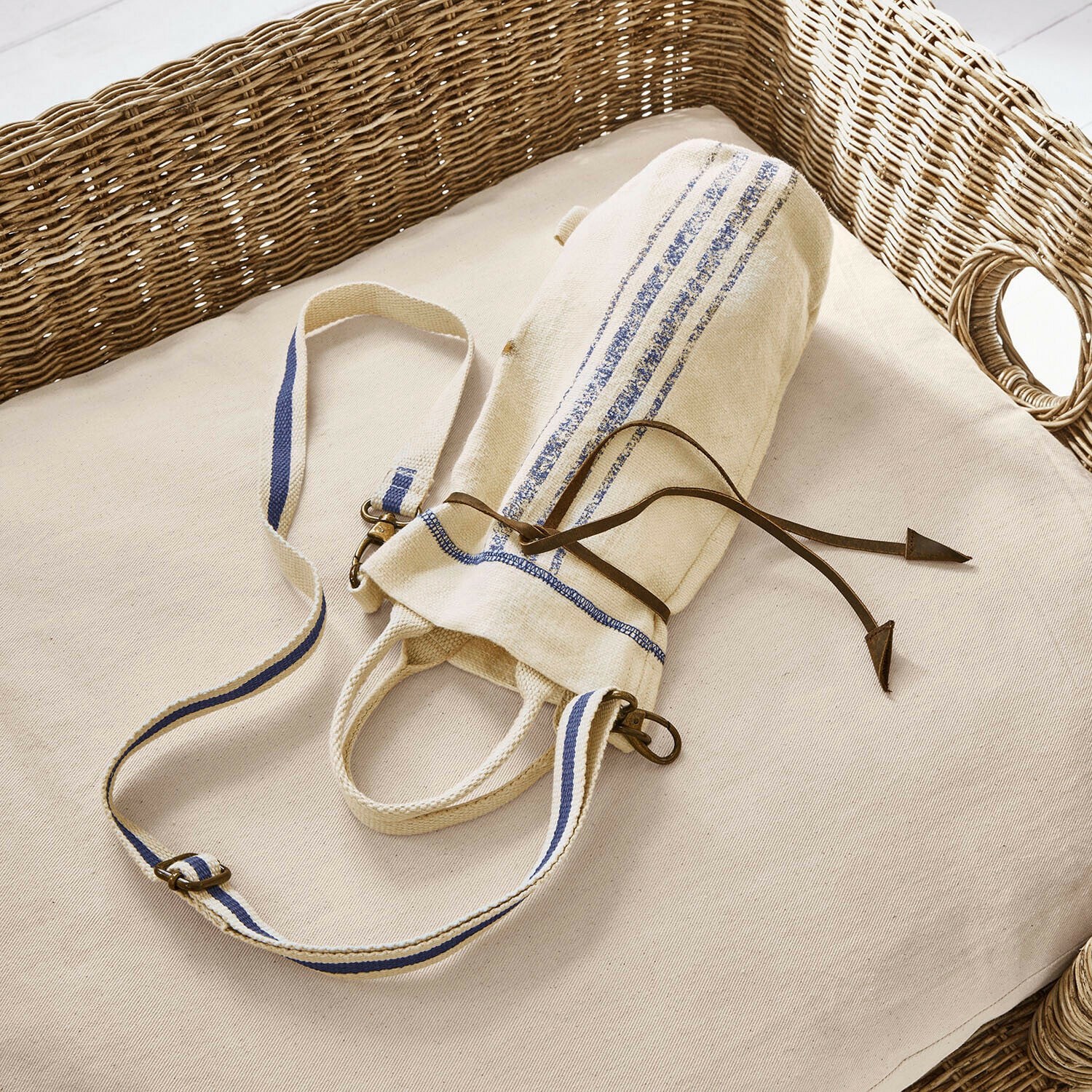 Flessentasje Pawera Beige katoenen tas met blauwe strepen en leren koord op licht kussen met rieten rugleuning