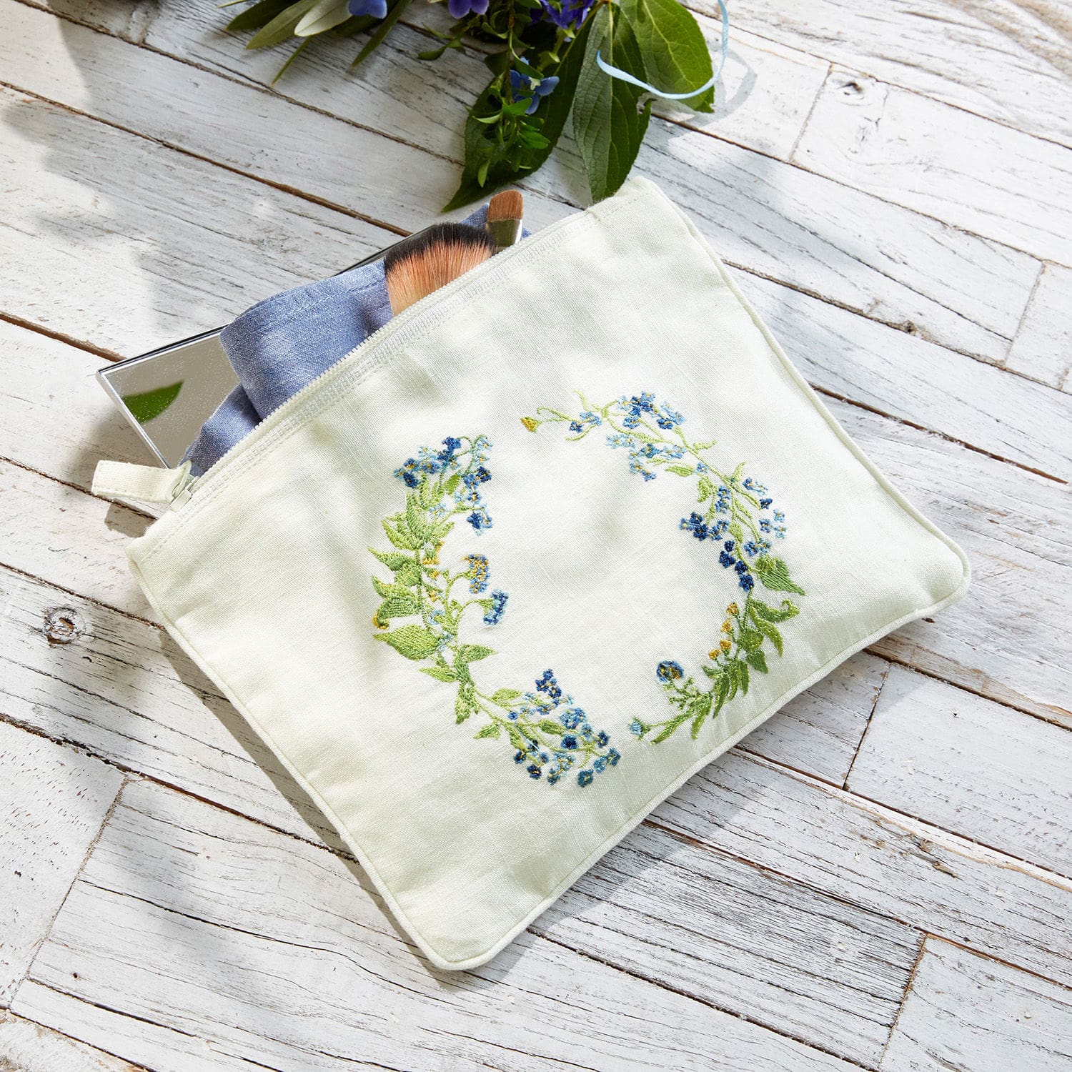 Trousse de maquillage en lin blanc avec broderie florale bleue posée sur un fond en bois clair