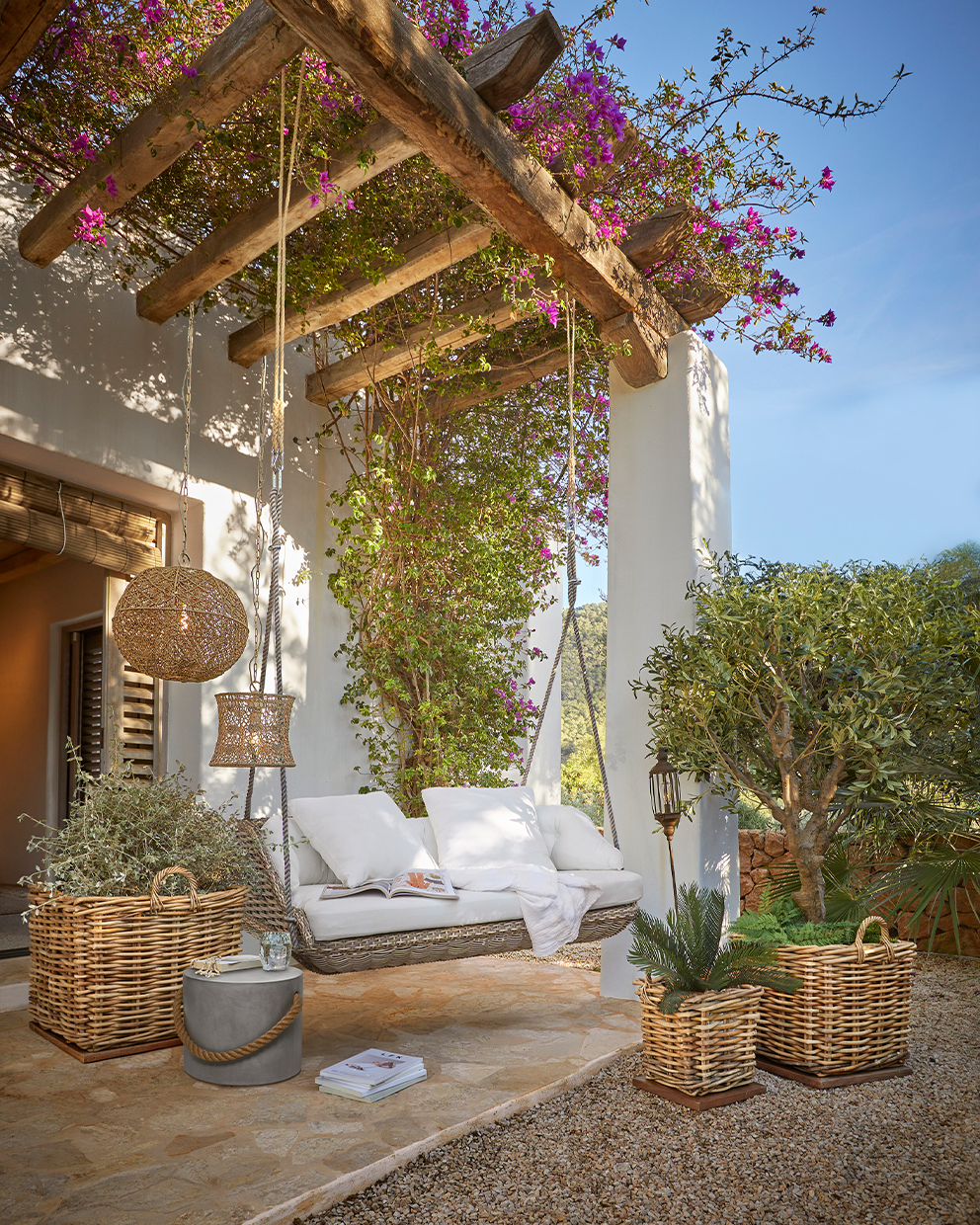 Rotan schommel met witte kussens onder een houten pergola met bloeiende roze bloemen