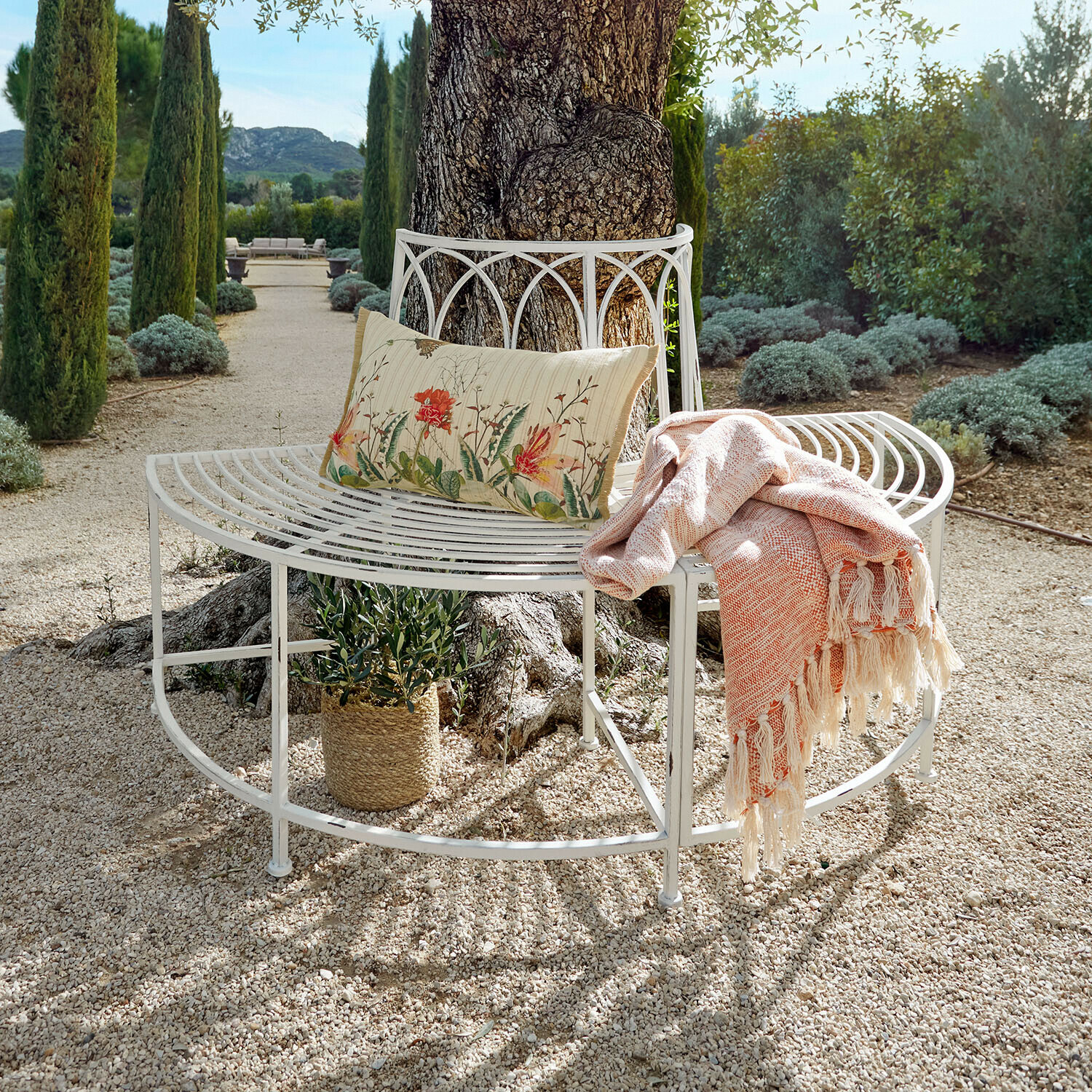 Banc Sherburne Banc de jardin en métal blanc autour d'un arbre avec coussin floral et plaid orange sur chemin de gravier