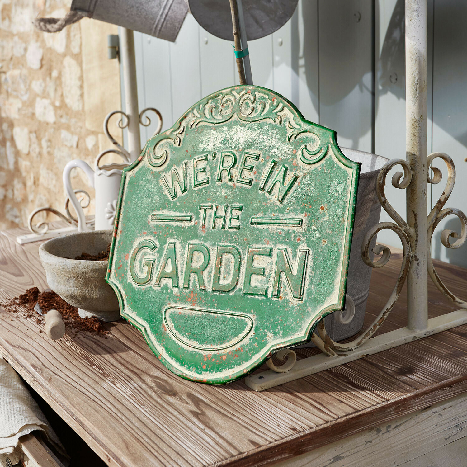 Panneau décoratif Merrin Plaque décorative en métal vert avec inscription We're in the Garden posée sur une table en bois dans un jardin