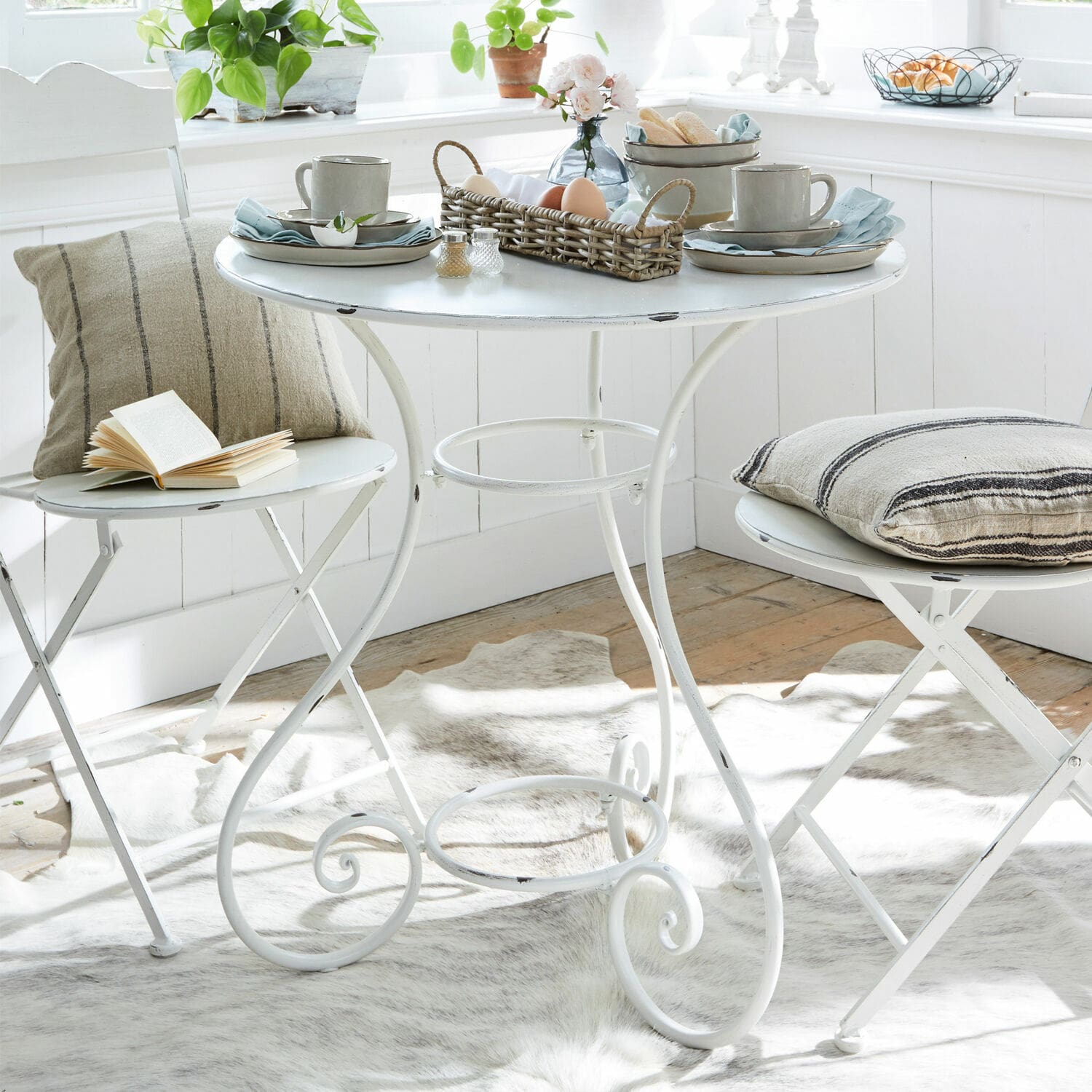 Table ronde en métal blanc avec deux chaises pliantes et coussins décoratifs dans un intérieur lumineux