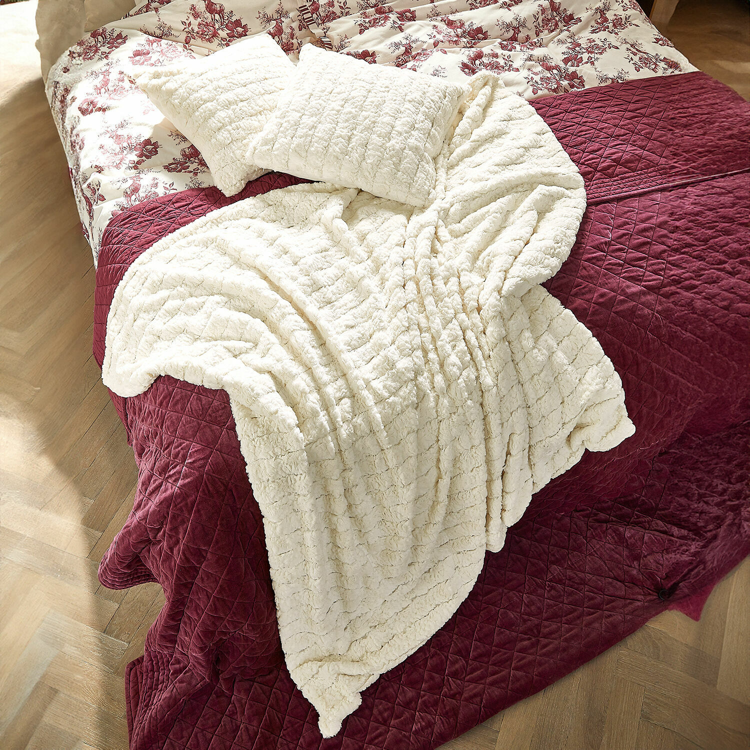 Plaid Dorelice Plaid et coussins en peluche crème sur une couverture matelassée bordeaux sur le lit