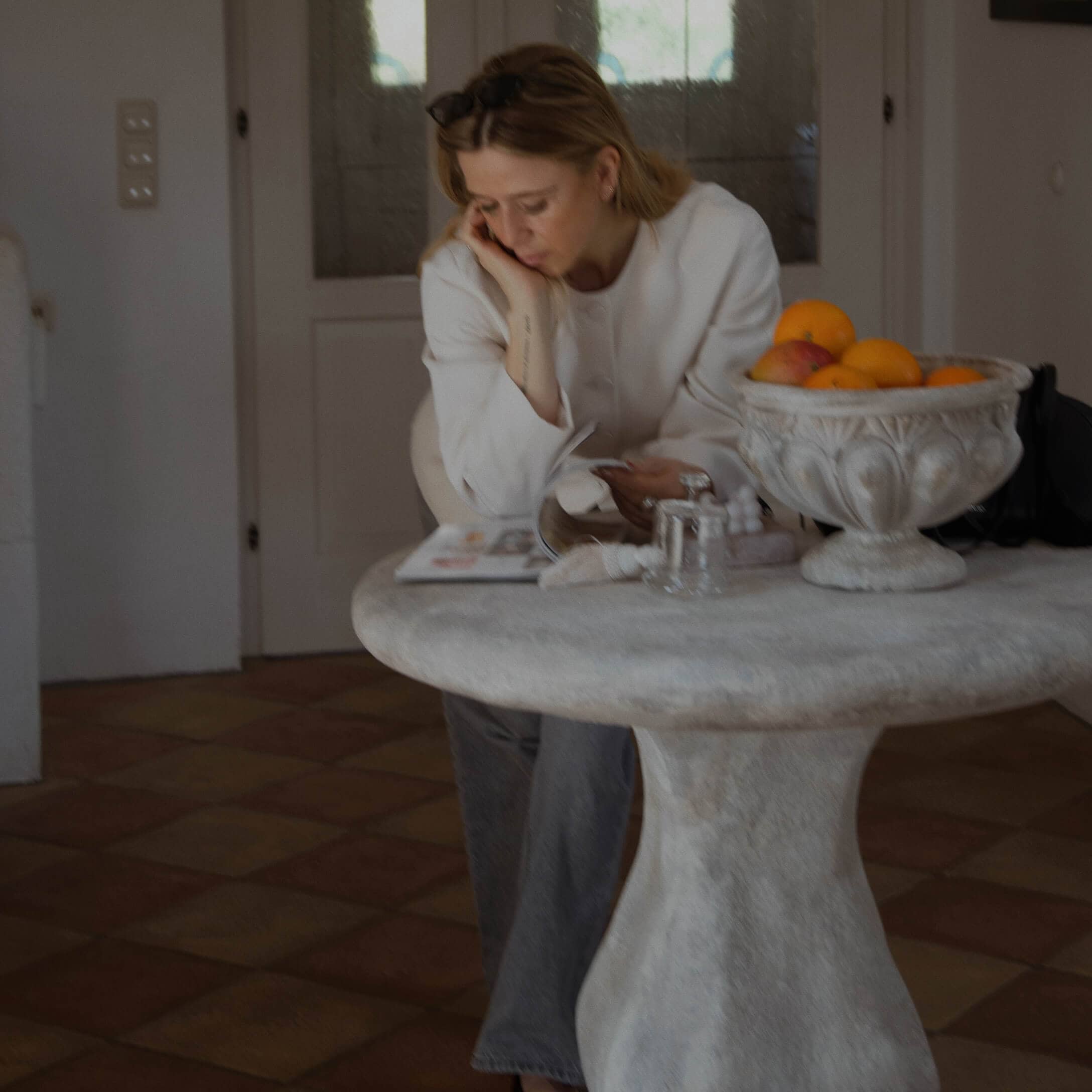 Femme en pull blanc lisant près d'une table ronde en pierre avec un bol de fruits à l'intérieur