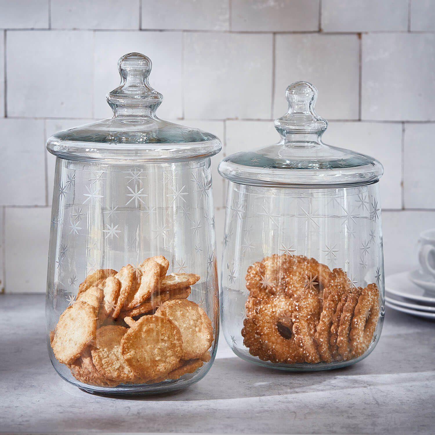 Twee doorzichtige glazen potten met deksels en sterpatroon, gevuld met goudbruine koekjes