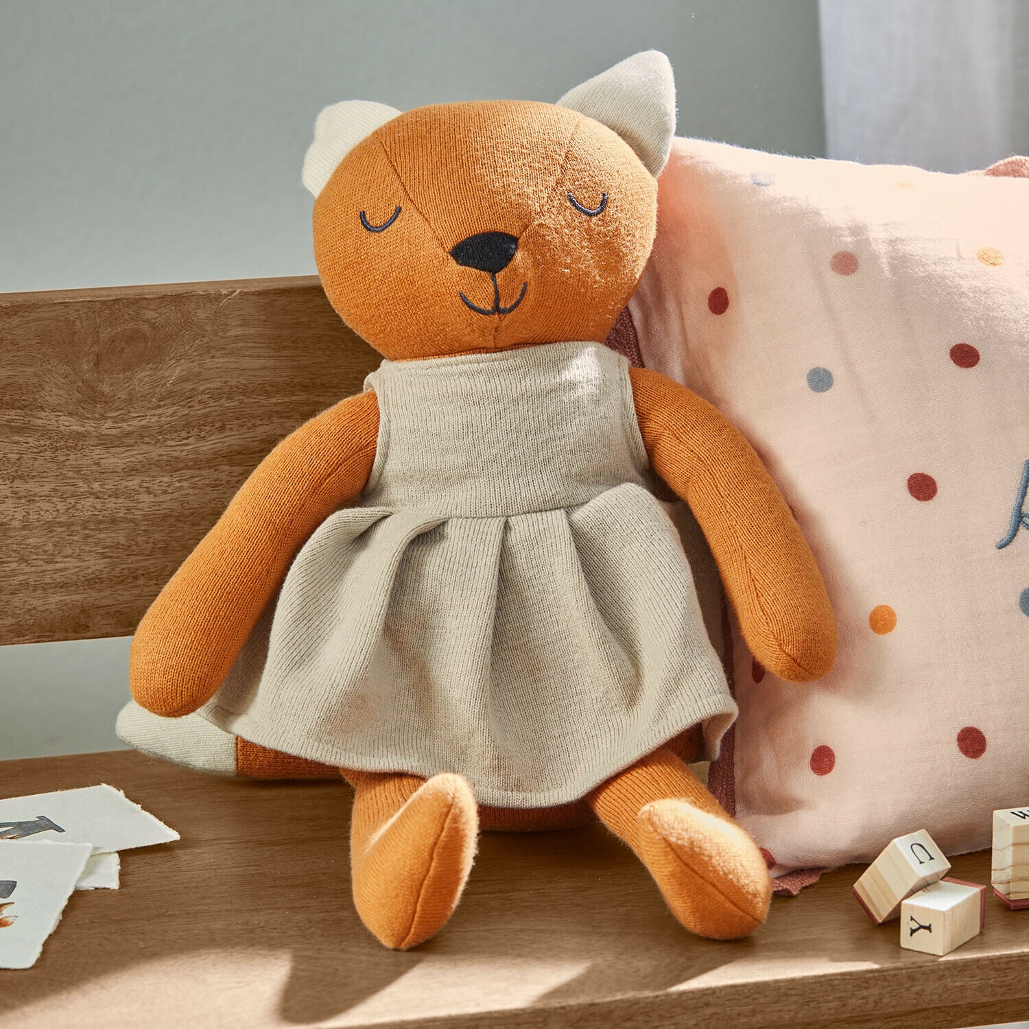 Peluche renard orange en robe beige assise sur un banc en bois avec coussin et cubes en bois