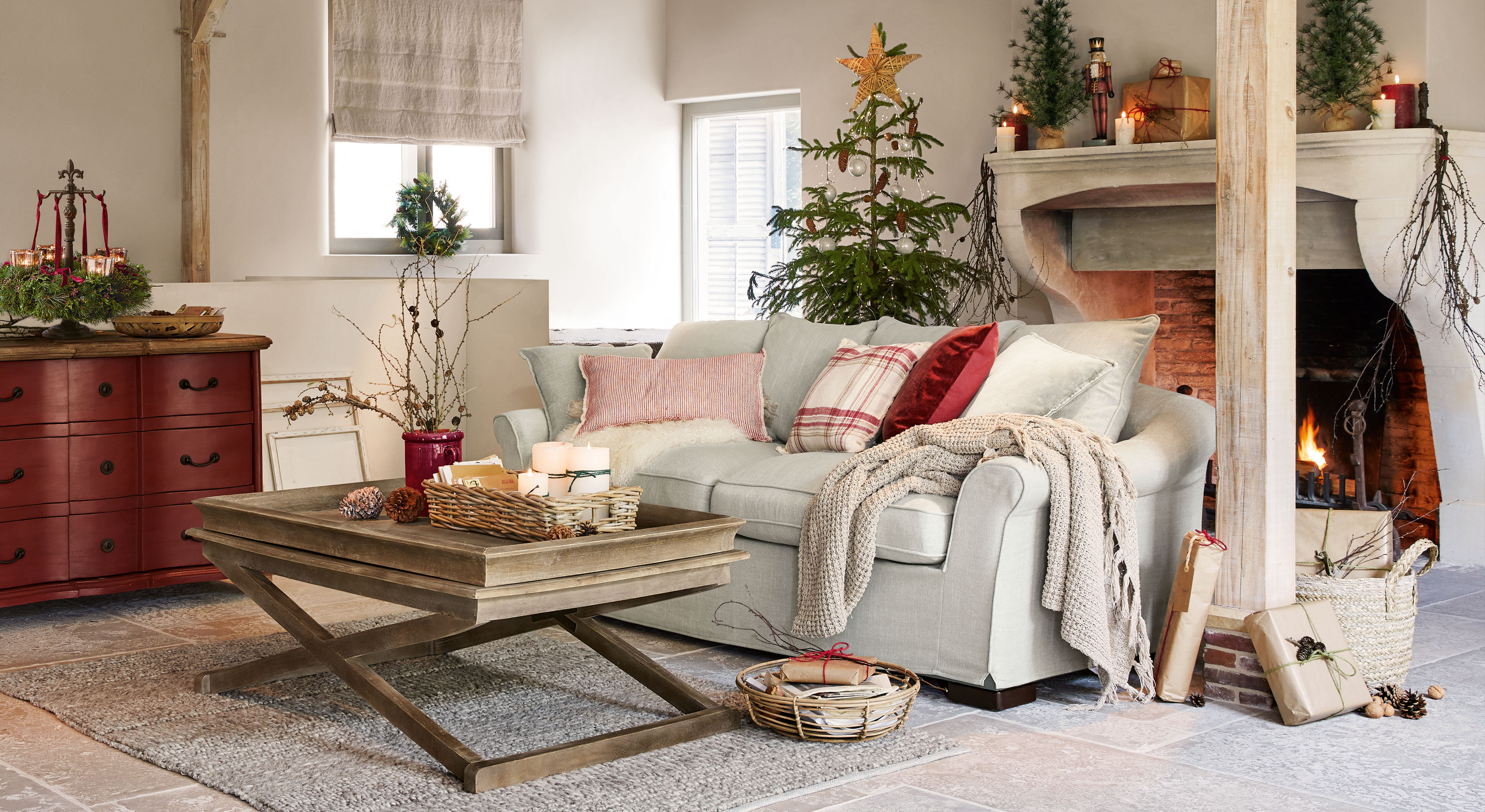 Salon chaleureux avec canapé gris clair, table basse en bois et décorations de Noël avec sapin et cheminée allumée