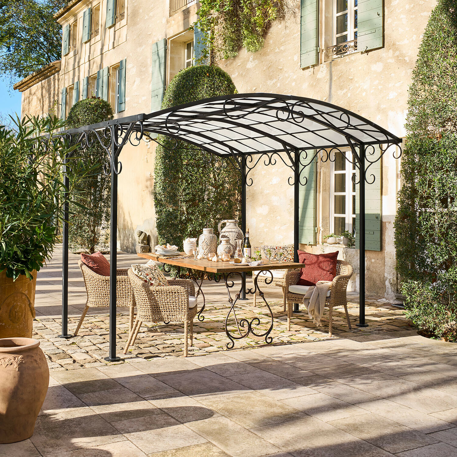 Pergola Delbridge Pergola en métal noir avec décorations en volutes au-dessus d'une table en bois et chaises en osier