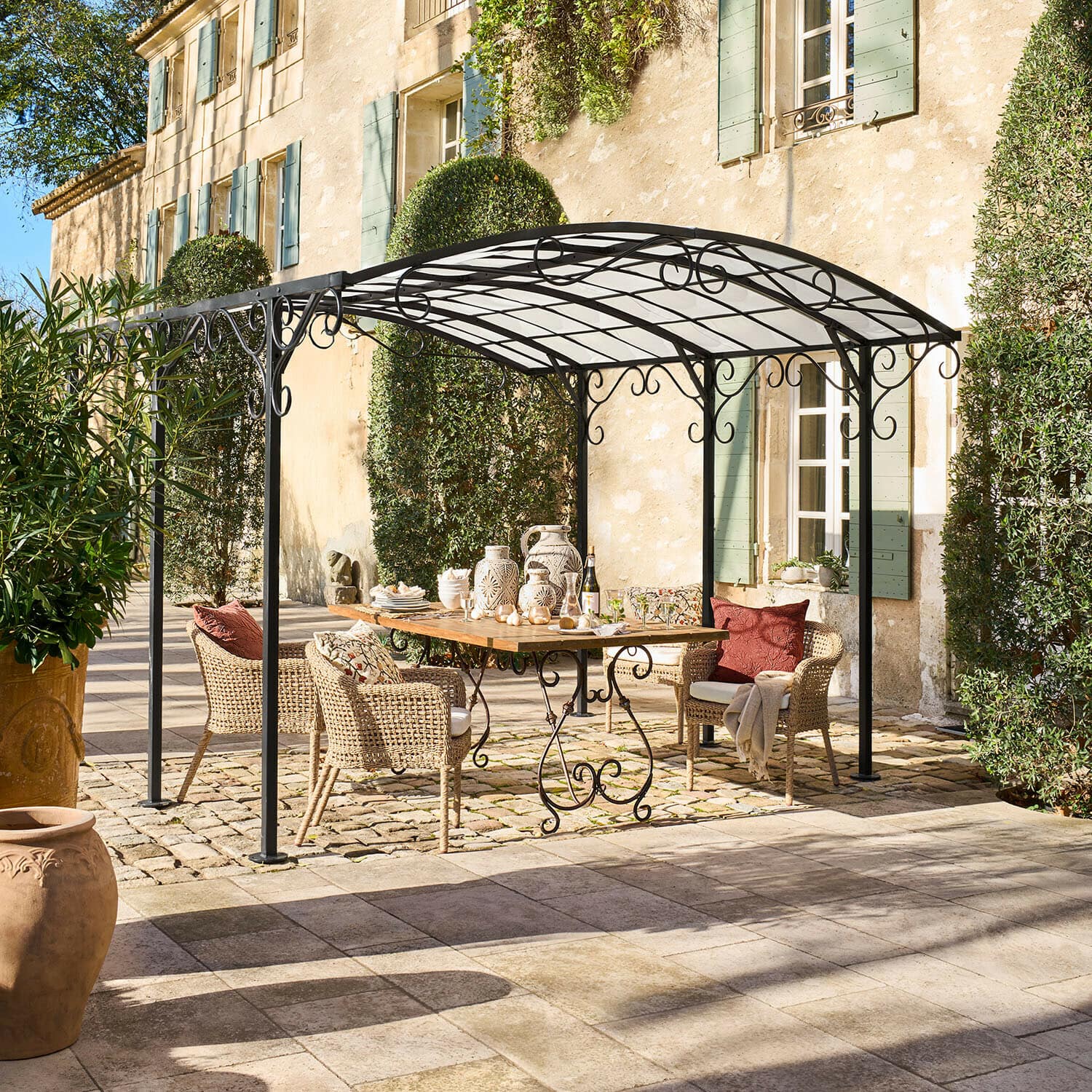 Pergola Delbridge Zwarte metalen tuinprieel met sierlijke krullen boven houten tafel en rotan stoelen op terras