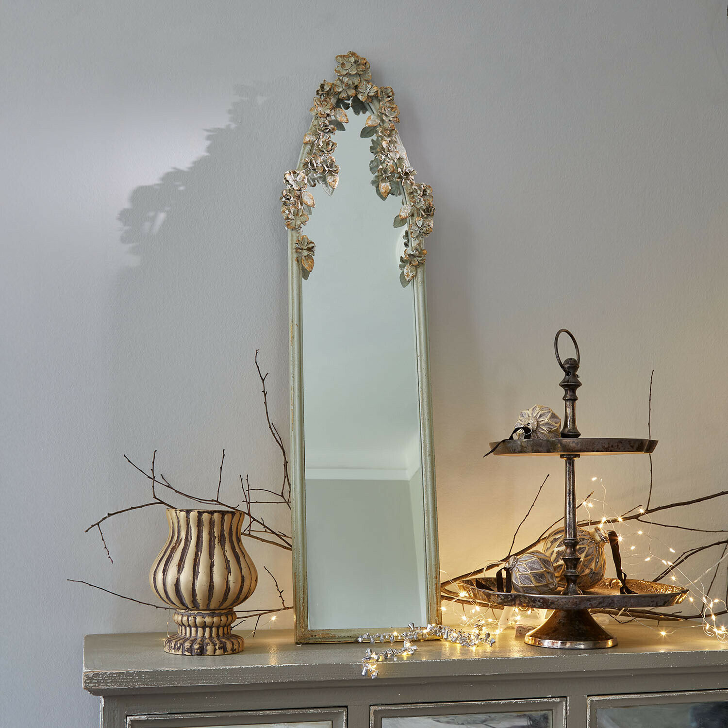 Miroir Clarendons Miroir étroit avec cadre doré vieilli et décor floral posé sur un meuble avec décorations lumineuses