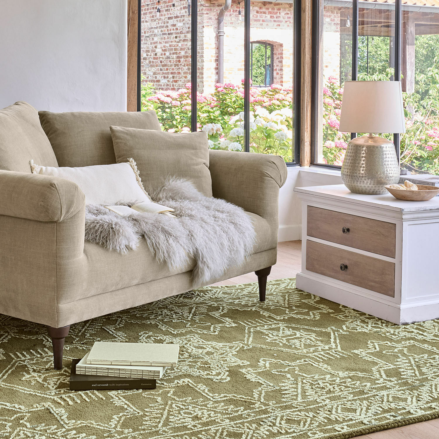 Vloerkleed Qazoura Beige stoffen bank met kussens en een bontdeken op een groen vloerkleed met geometrisch patroon
