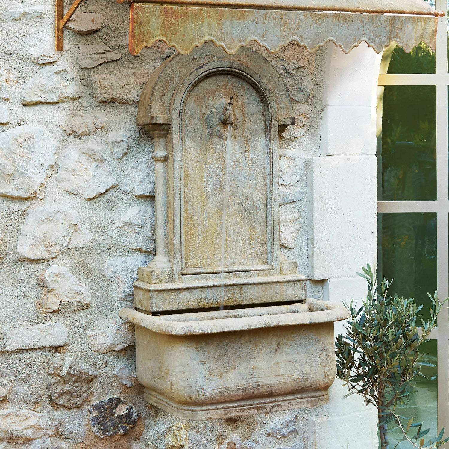 Muurbron Ivera Decoratieve beige stenen wandfontein met roestige metalen kap en stromend water buiten