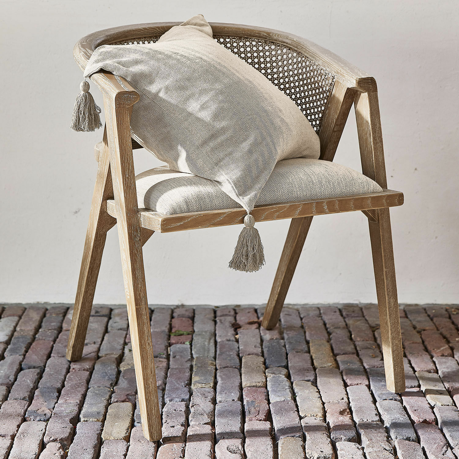 Stoel Collaroy Stoel van licht hout met gevlochten rugleuning en beige kussen met kwastjes op stenen vloer