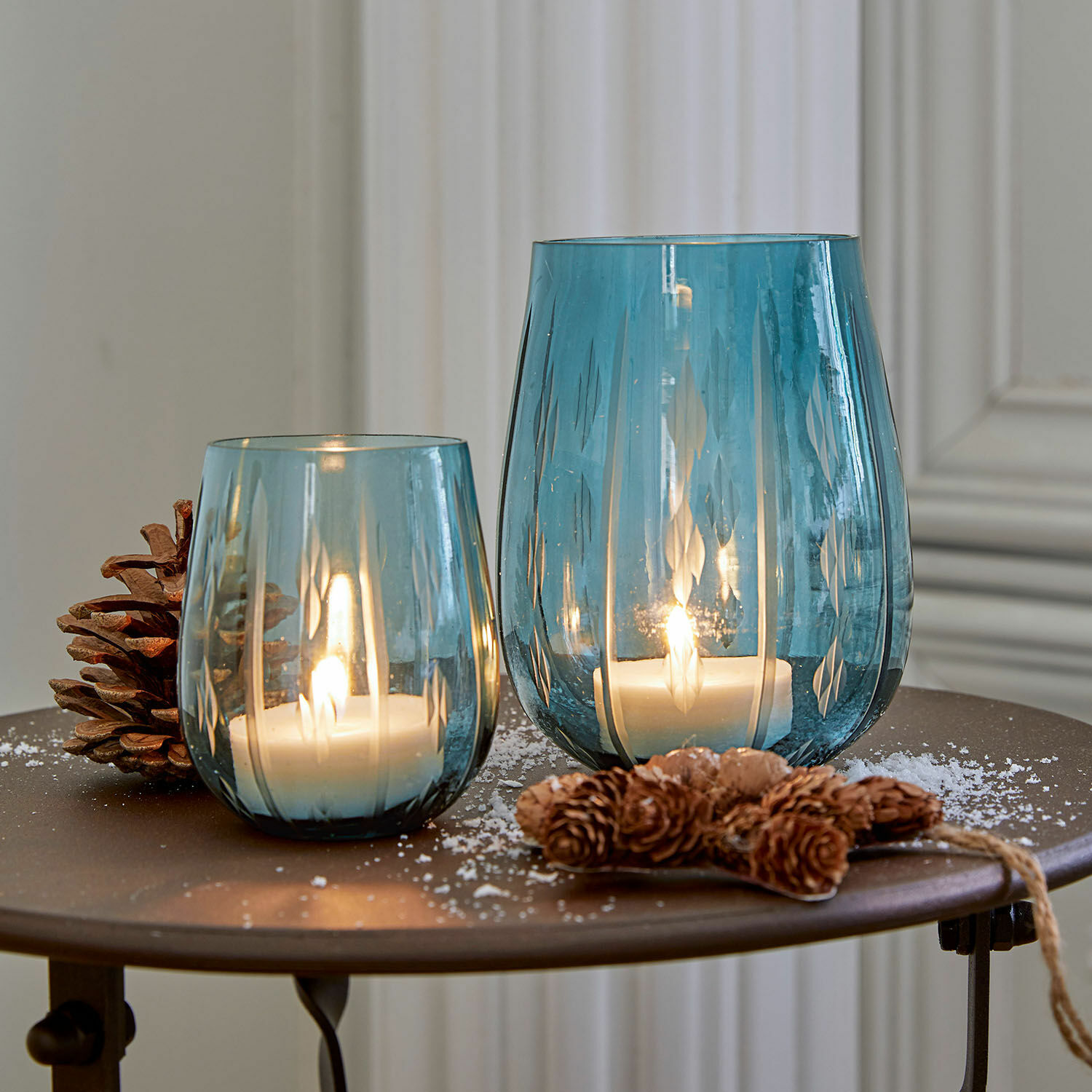Deux photophores en verre bleu avec motifs, bougies blanches allumées et décoration de pommes de pin