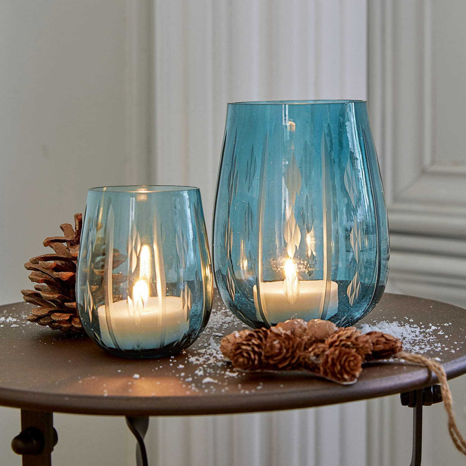 Lot de 2 photophores Danisar Deux photophores en verre bleu avec motifs, bougies blanches allumées et décoration de pommes de pin