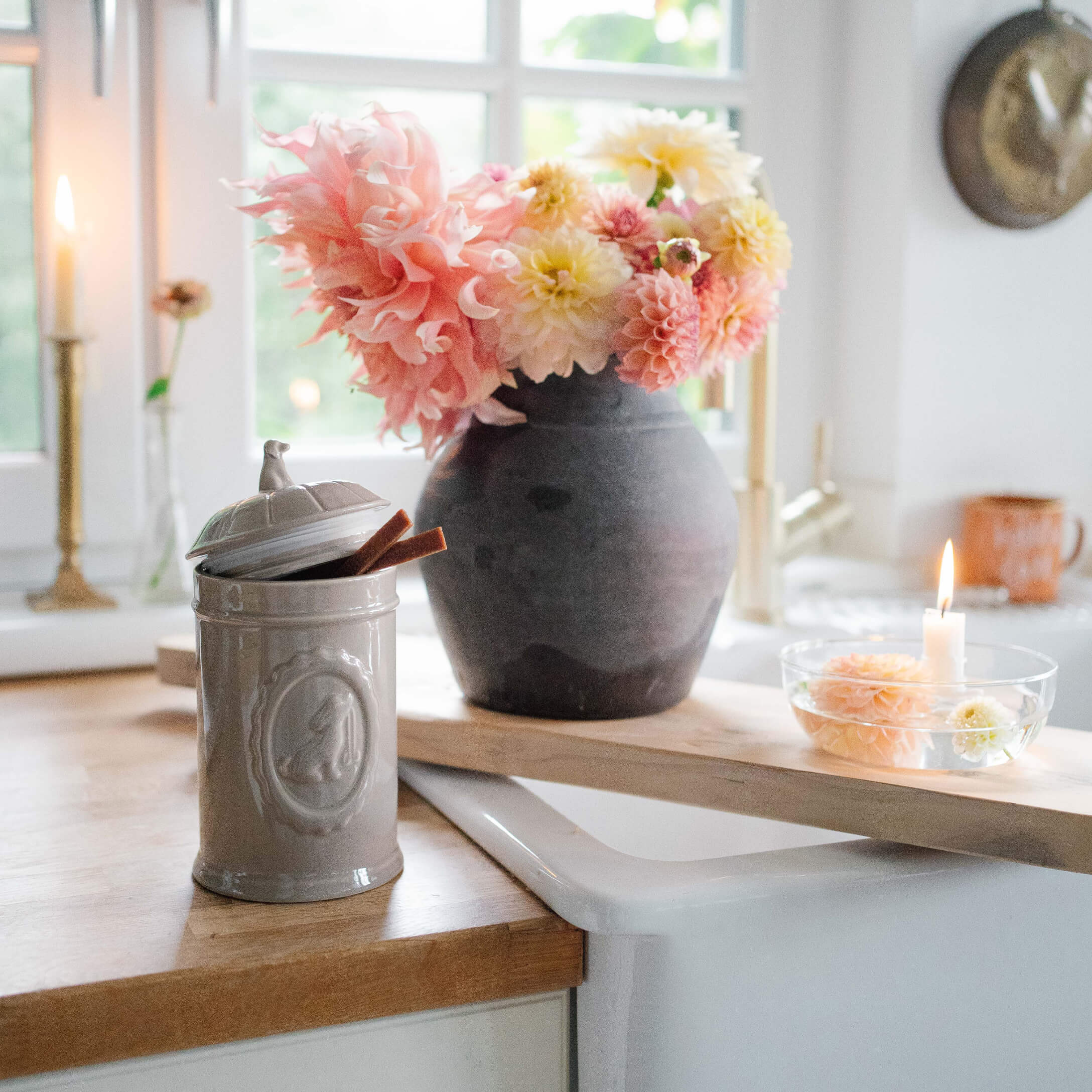 Grijze keramische pot met deksel op houten aanrecht naast zwarte vaas met roze en gele bloemen