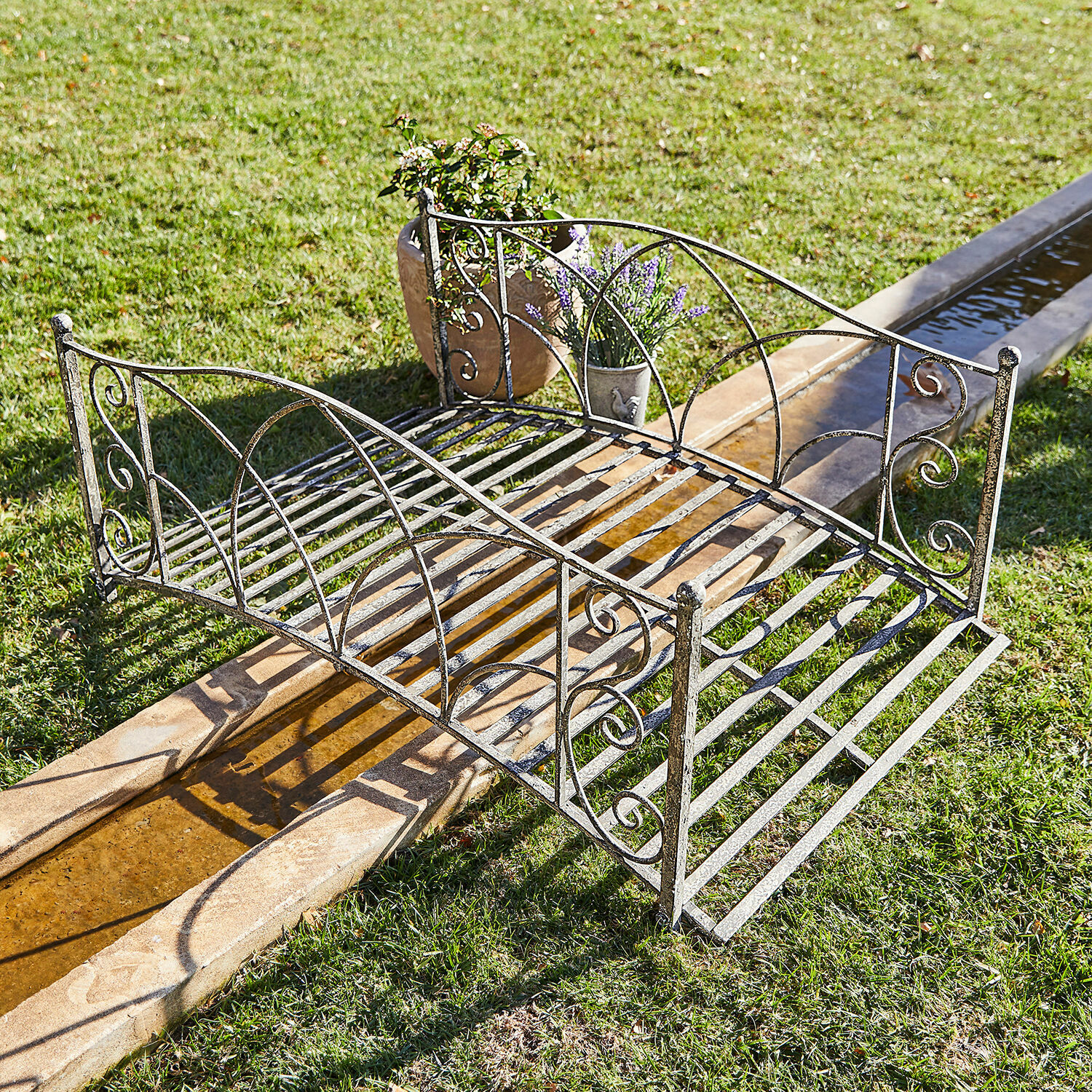 Decoratieve brug Escàs Sierlijke metalen tuinbrug in verouderd zilver over een waterloop op het gras met bloempotten