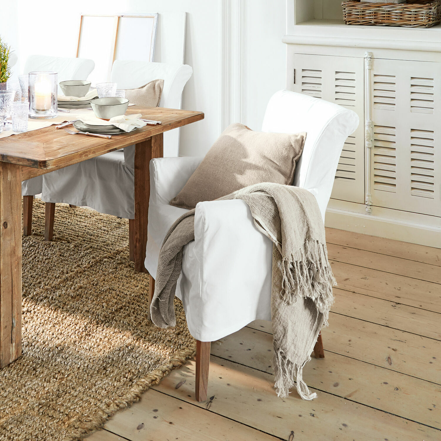 Chaise Amherst Fauteuil blanc avec pieds en bois, coussin beige et plaid naturel dans une salle à manger avec table en bois