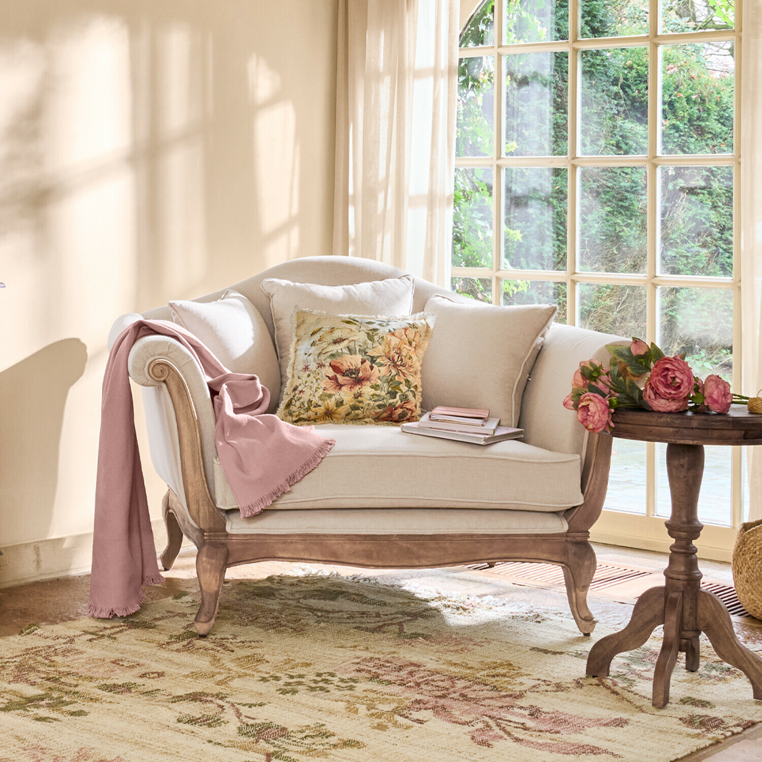 Canapé beige en bois avec coussin floral et plaid rose dans un salon lumineux avec grande fenêtre