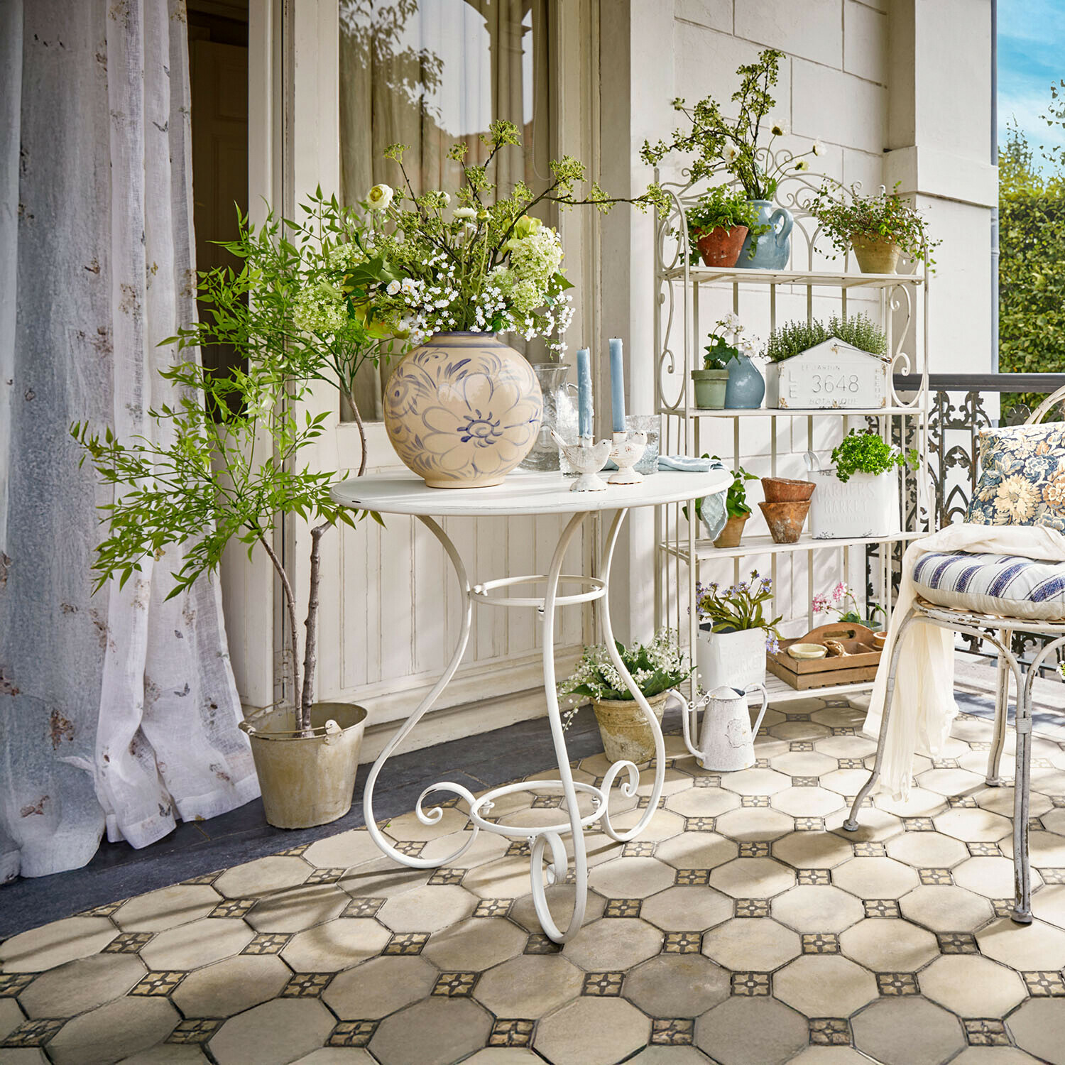 Witte metalen tafel met decoratieve vaas en bloemen op terras met planten en stoel