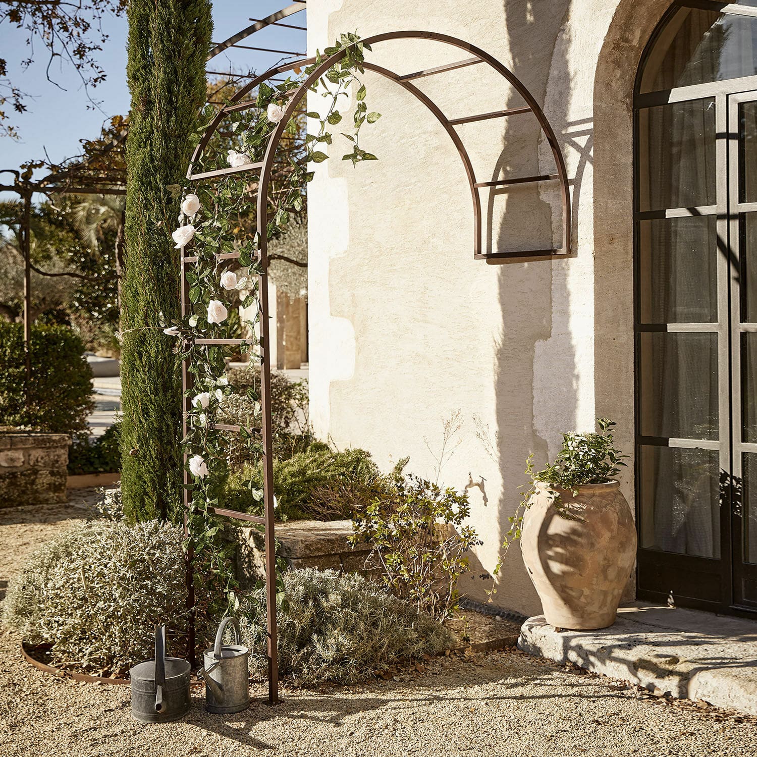 Bruine metalen tuinpergola met boog, witte rozen en gieters op grind