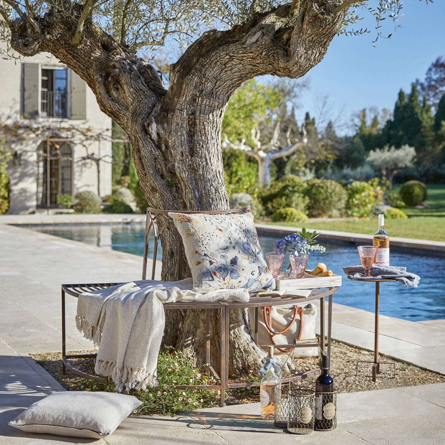 Banc Sherburne Banc en métal autour d'un arbre avec coussin et plaid près d'une piscine dans un jardin ensoleillé