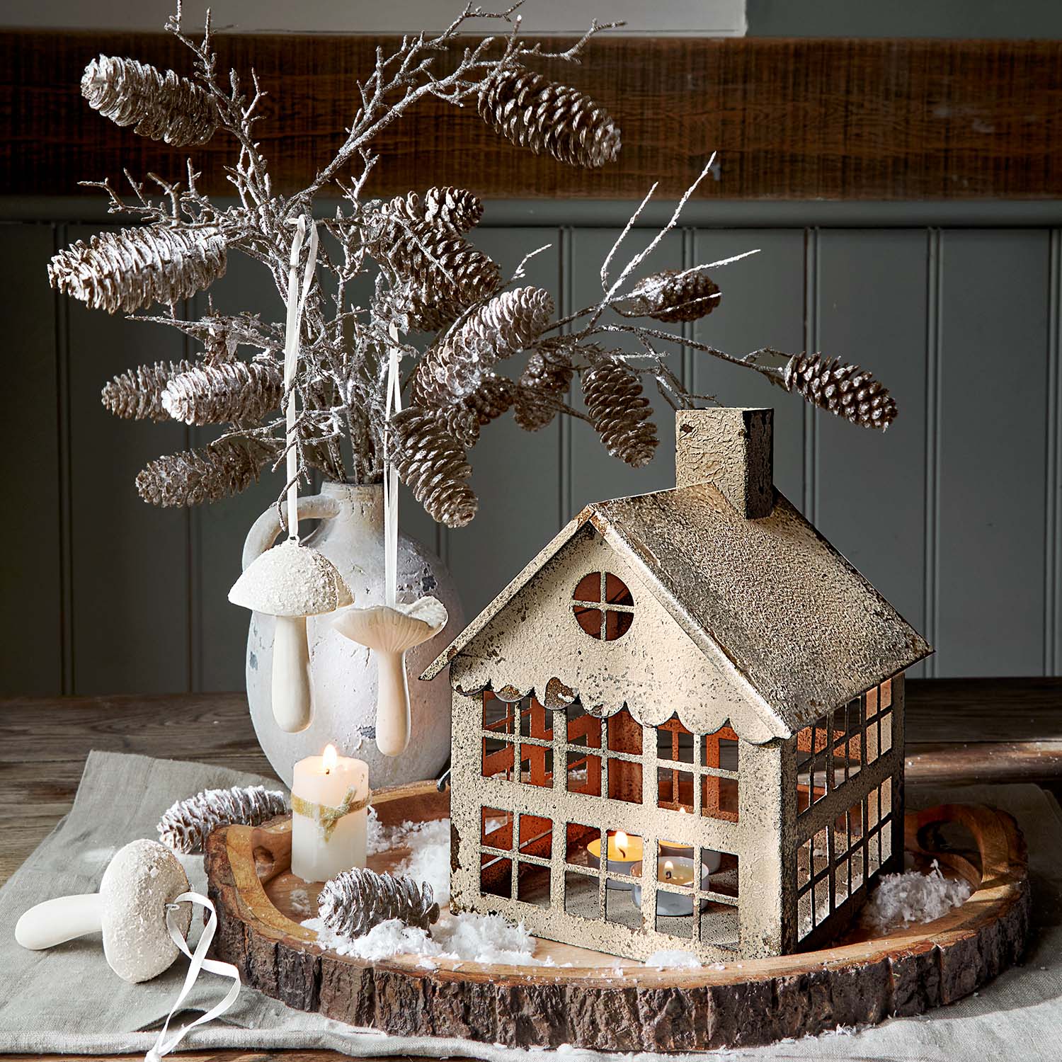 Lanterne décorative en métal beige en forme de maison avec branches de pommes de pin et bougie sur plateau en bois