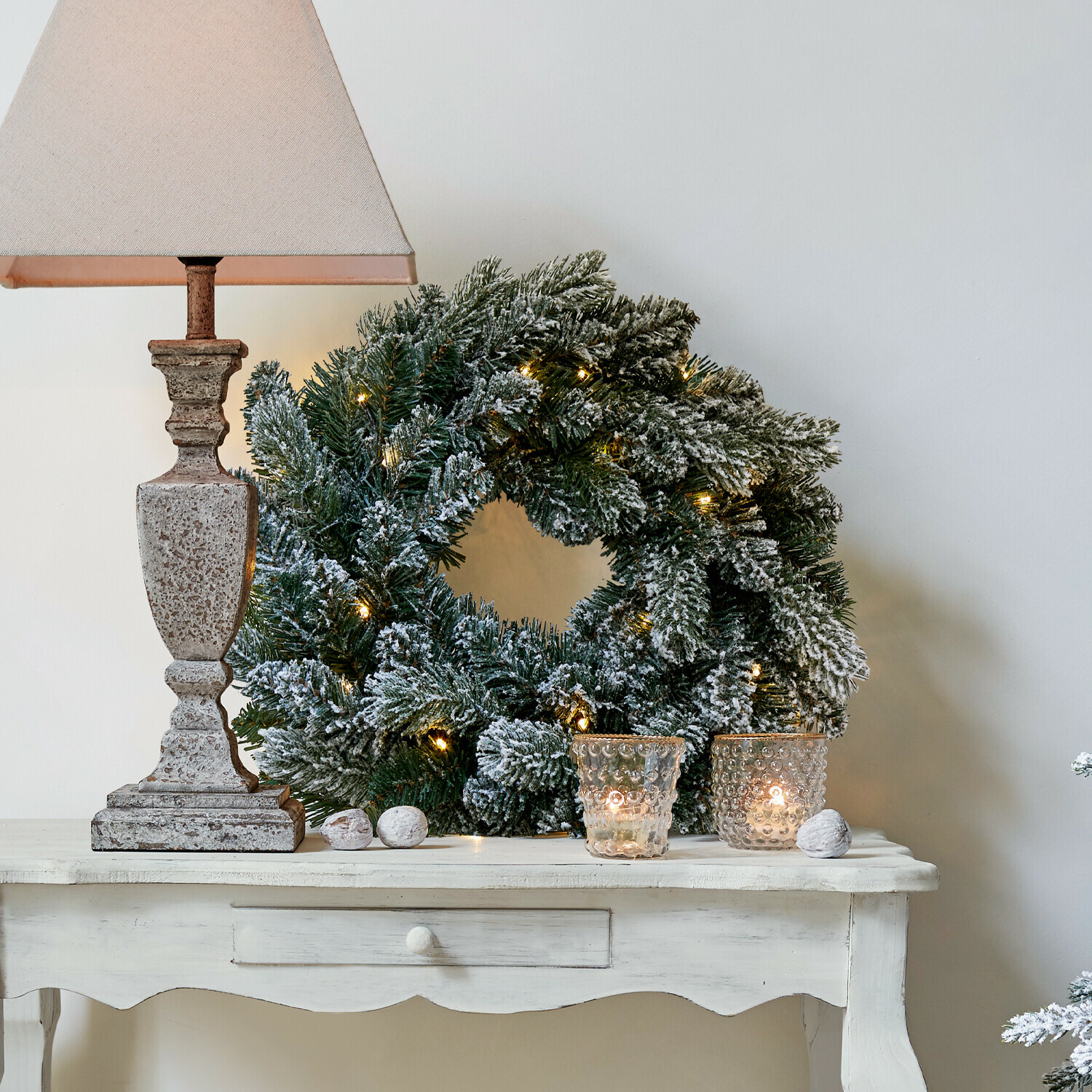 Couronne LED Regin Couronne de Noël enneigée avec branches vertes et lumières sur une table blanche avec lampe