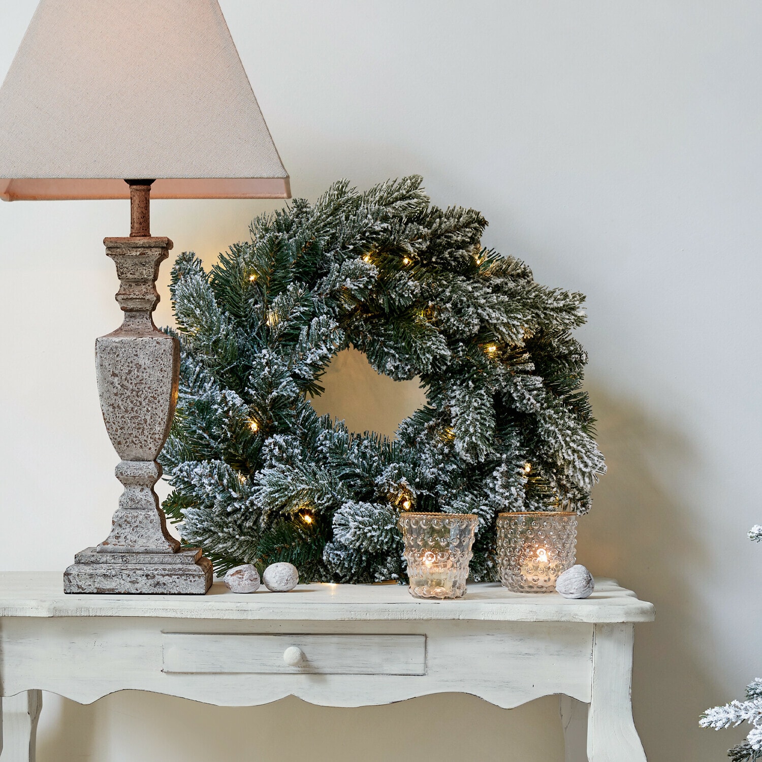 Couronne LED Regin Couronne de Noël enneigée avec branches vertes et lumières sur une table blanche avec lampe