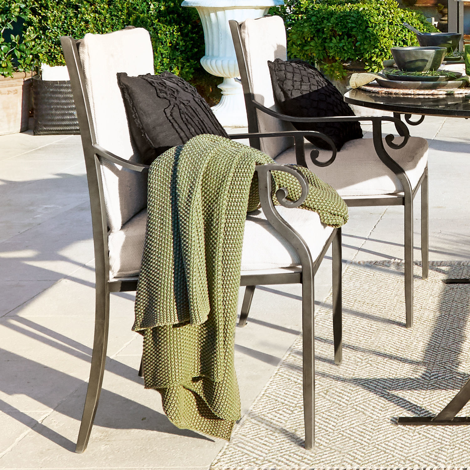 Plaid Amberley Metalen tuinstoelen met crèmekleurige kussens en groene gebreide deken op lichte stenen terras