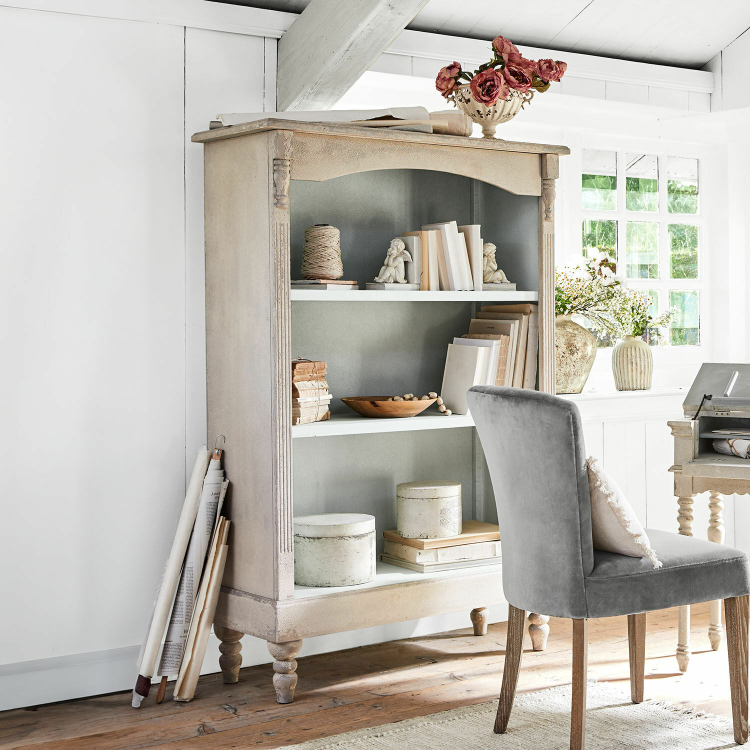 Étagère Arterton Bibliothèque vintage en bois beige avec livres décoratifs et chaise grise dans une pièce lumineuse