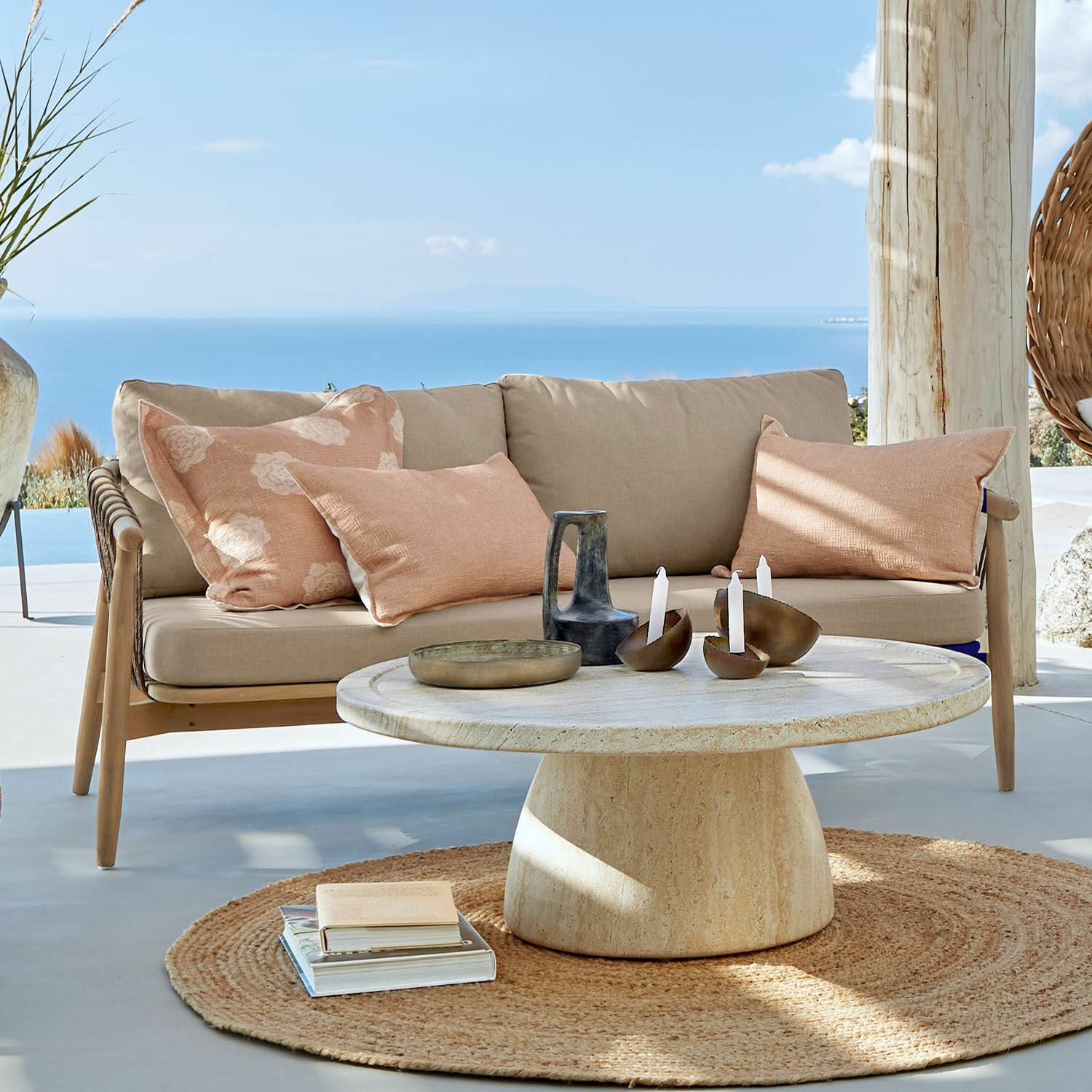 Bank FSC®-100% Thornwick Beige bank met kussens en ronde stenen tafel op terras met zeezicht