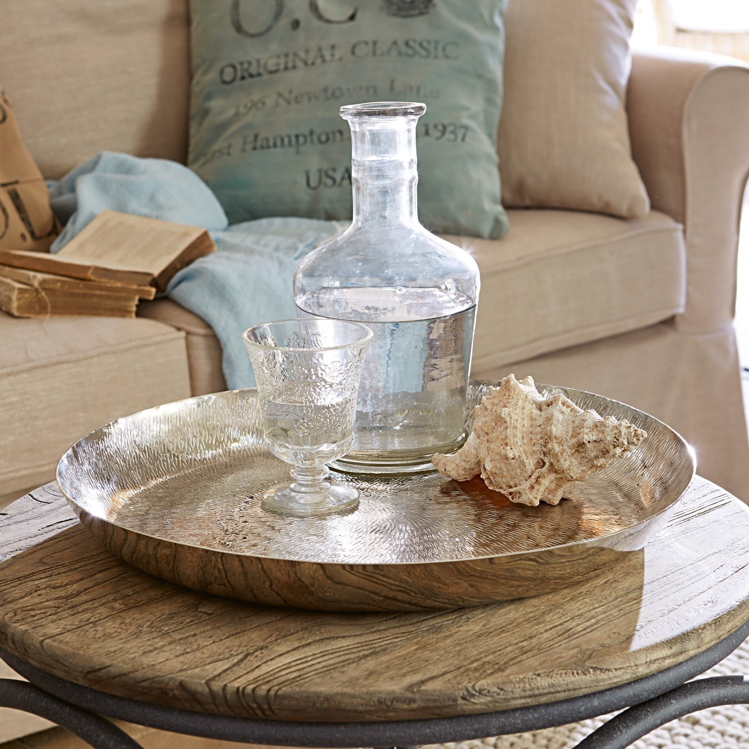 Plateau Caelan Plateau rond en métal argenté sur table en bois avec carafe en verre et coquillage décoratif