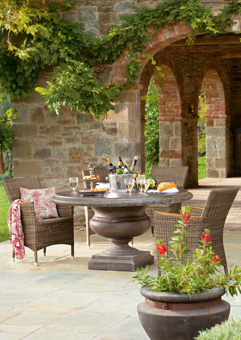 Table de jardin ronde en pierre avec chaises en osier sur patio en pierre et arches en pierre avec verdure