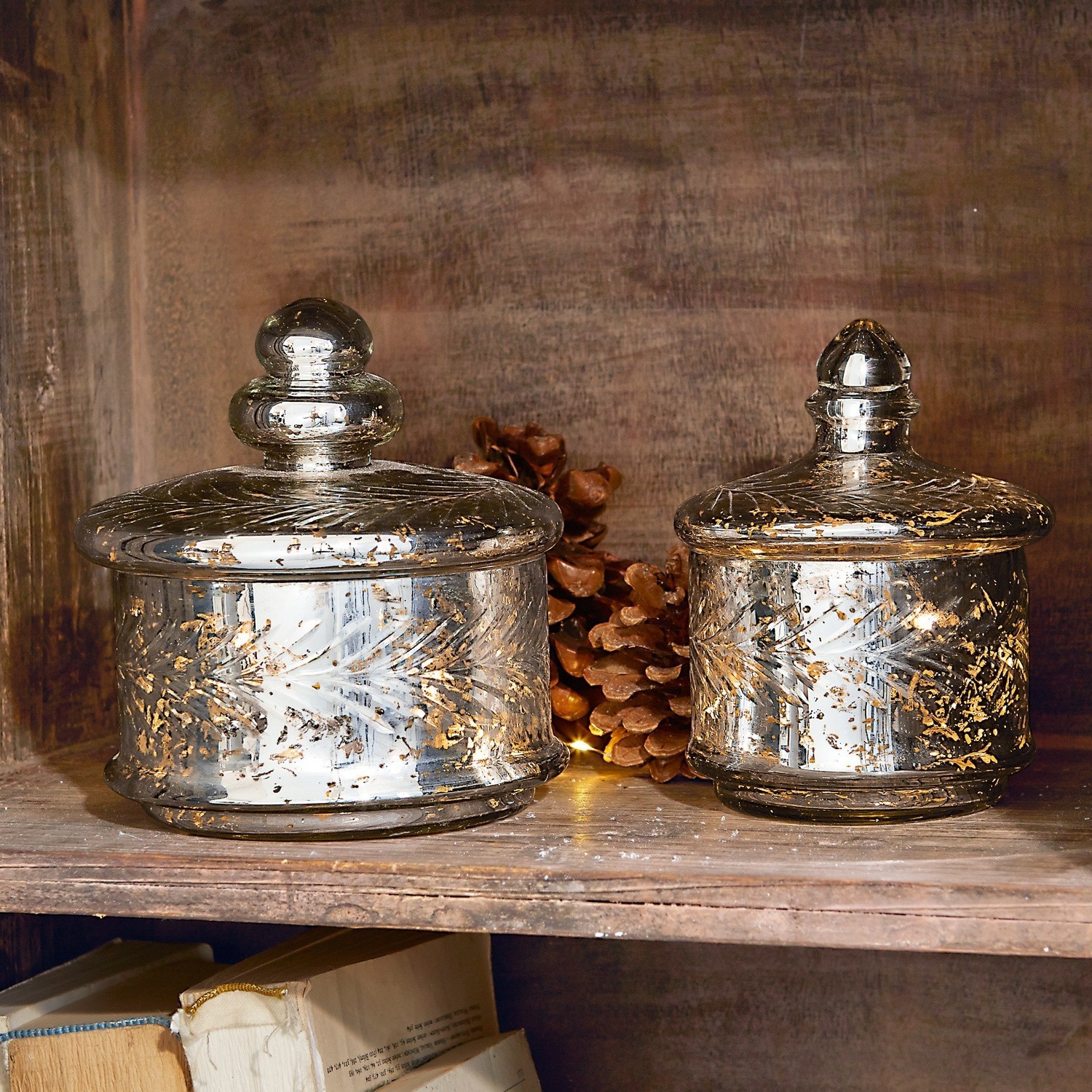 Glazen pot set van 2 Elexa Twee glazen potten met deksels en gouden versieringen op houten plank met dennenappels