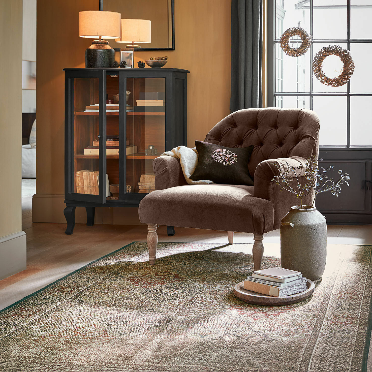 Vloerkleed voor binnen en buiten Malhira Bruine fluwelen fauteuil met gestikte rugleuning in gezellige woonkamer met tapijt en zwarte vitrine
