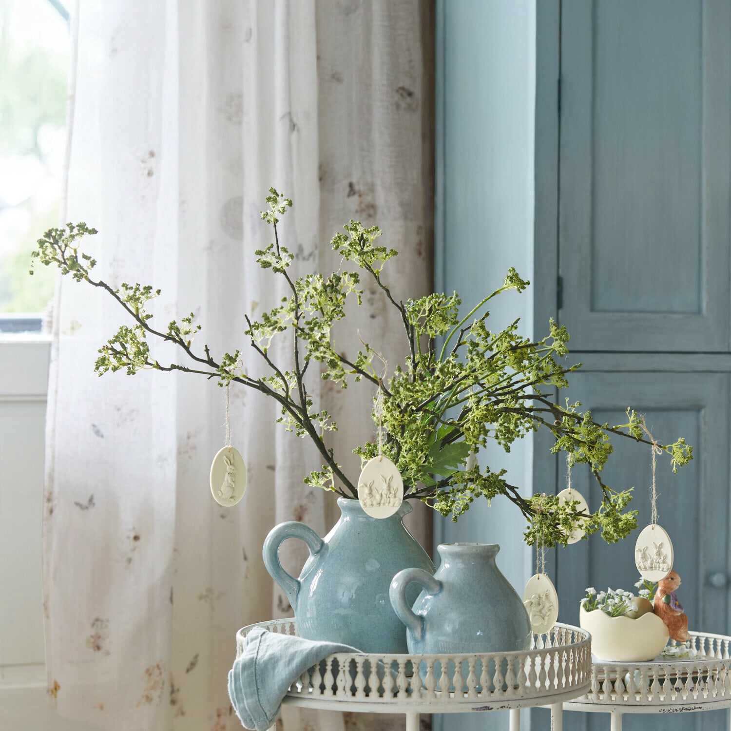 Blauwe keramische kannen met groene takken en paasdecoraties op een witte metalen tafel