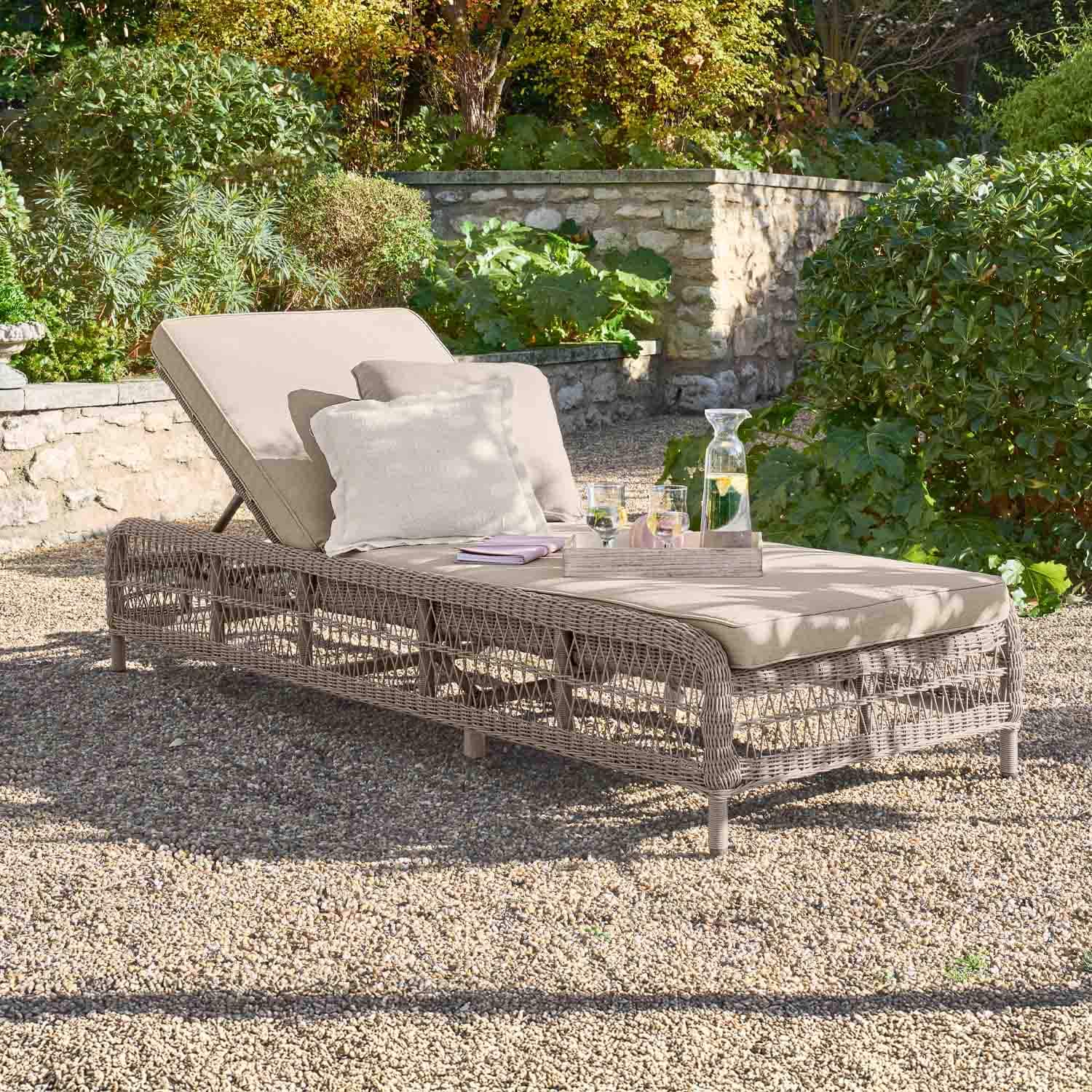 Chaise longue de jardin beige avec coussin et structure en osier sur sol gravillonné dans un jardin verdoyant