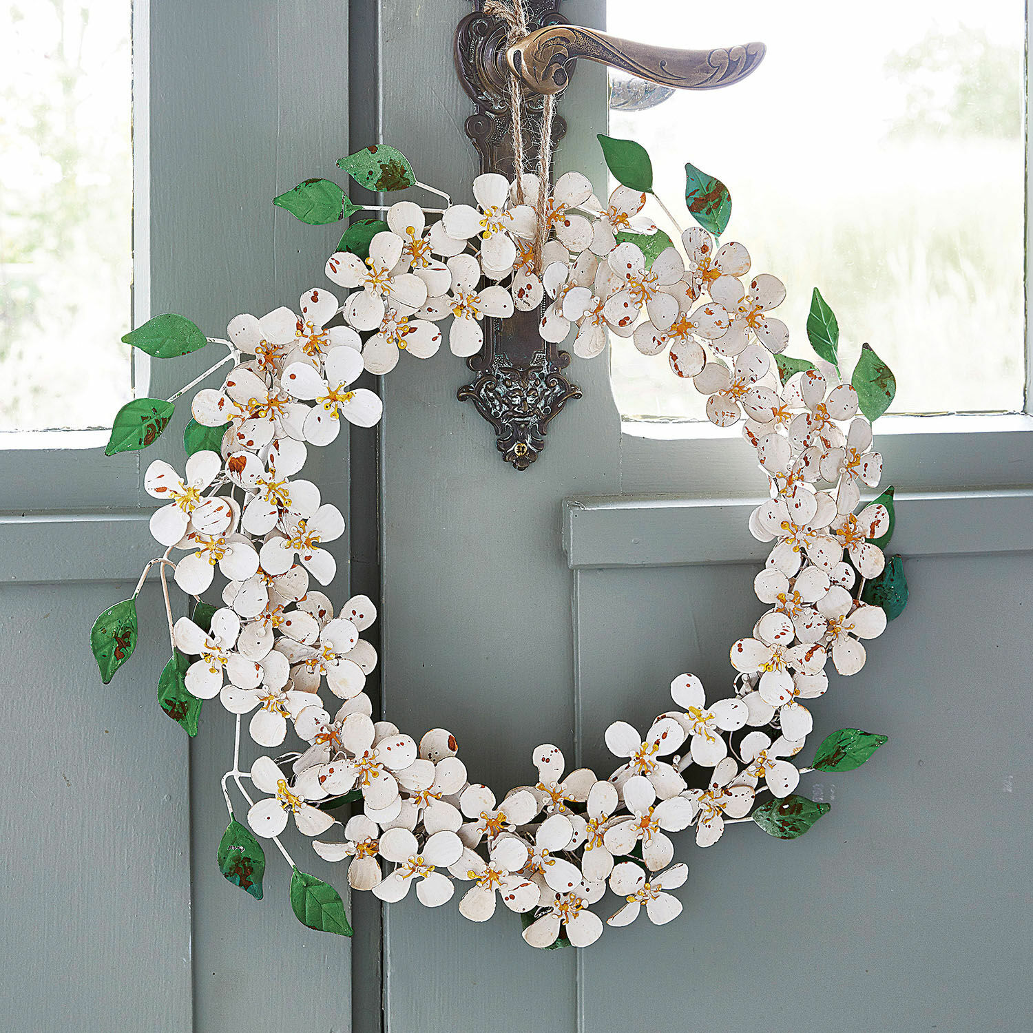 Couronne Loanne Couronne décorative en métal blanc avec fleurs et feuilles vertes suspendue à une porte grise