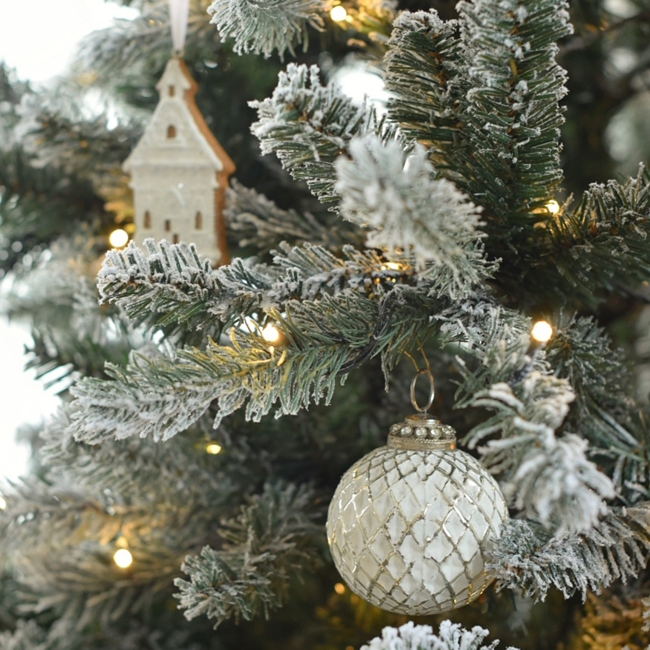 Witte glazen kerstbal met ruitpatroon aan besneeuwde kerstboom met LED-lichtjes en houten decoratie