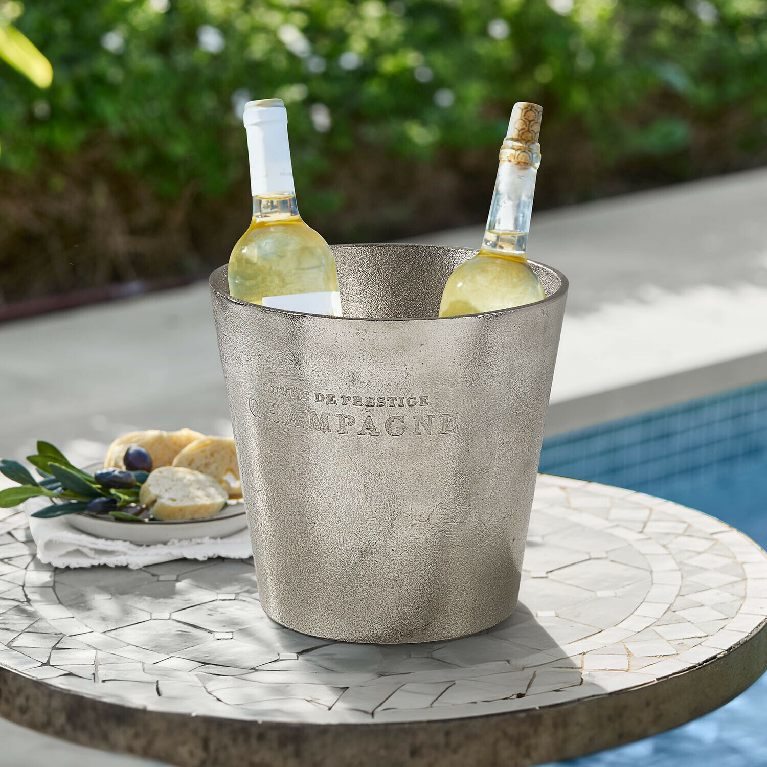 Seau à glace en métal argenté avec inscription Champagne contenant deux bouteilles de vin blanc sur une table extérieure