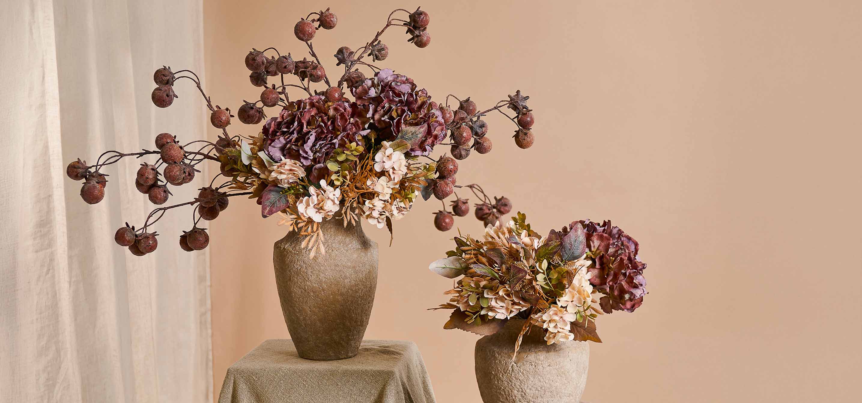 Twee decoratieve vazen van beige steen met boeketten van kunstbloemen en vruchten in warme kleuren