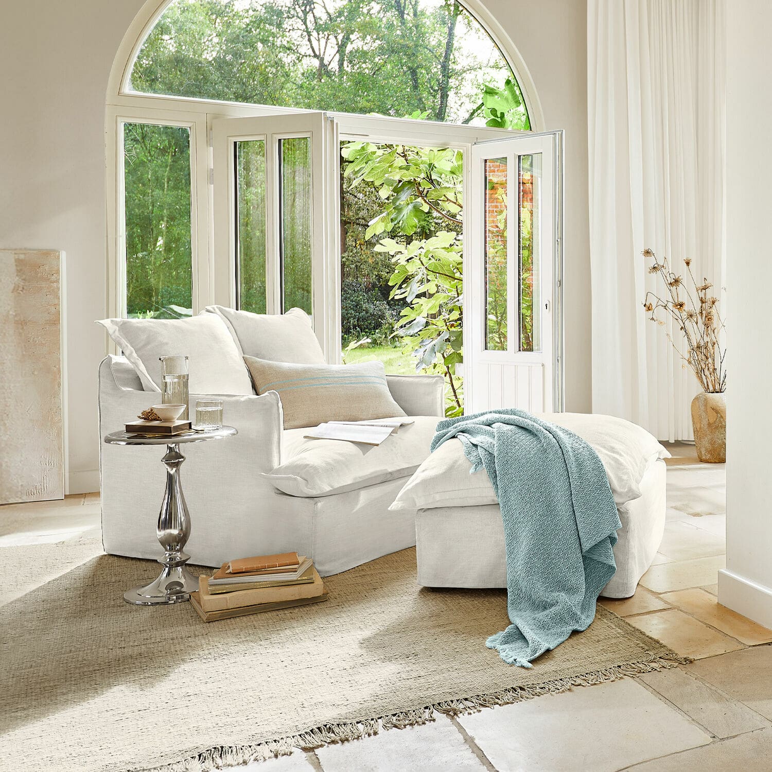 Fauteuil avec repose-pieds Avignon Fauteuil en coton blanc avec repose-pieds et couverture bleue dans un salon lumineux avec grande fenêtre