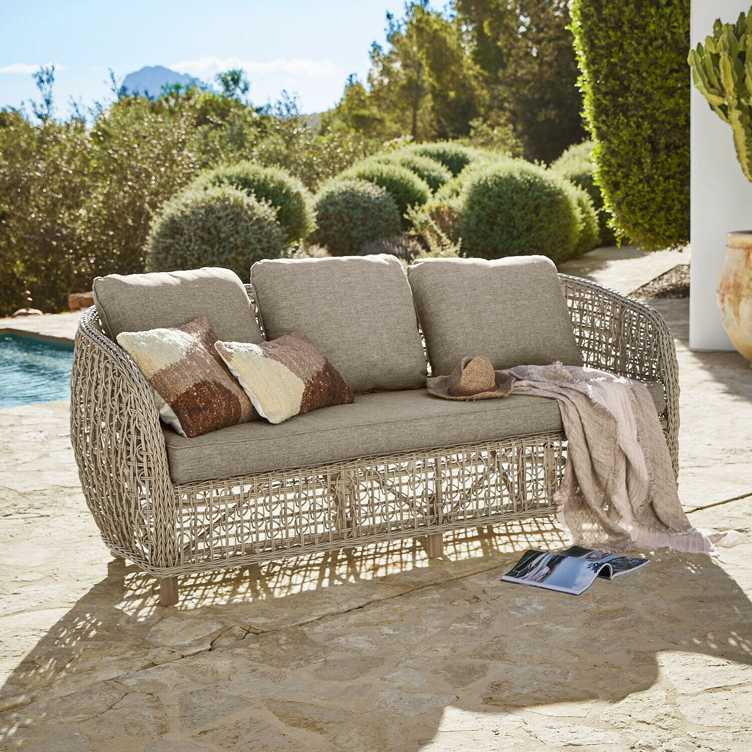 Canapé d'extérieur beige avec coussins et structure en rotin tressé sur terrasse en pierre près de la piscine